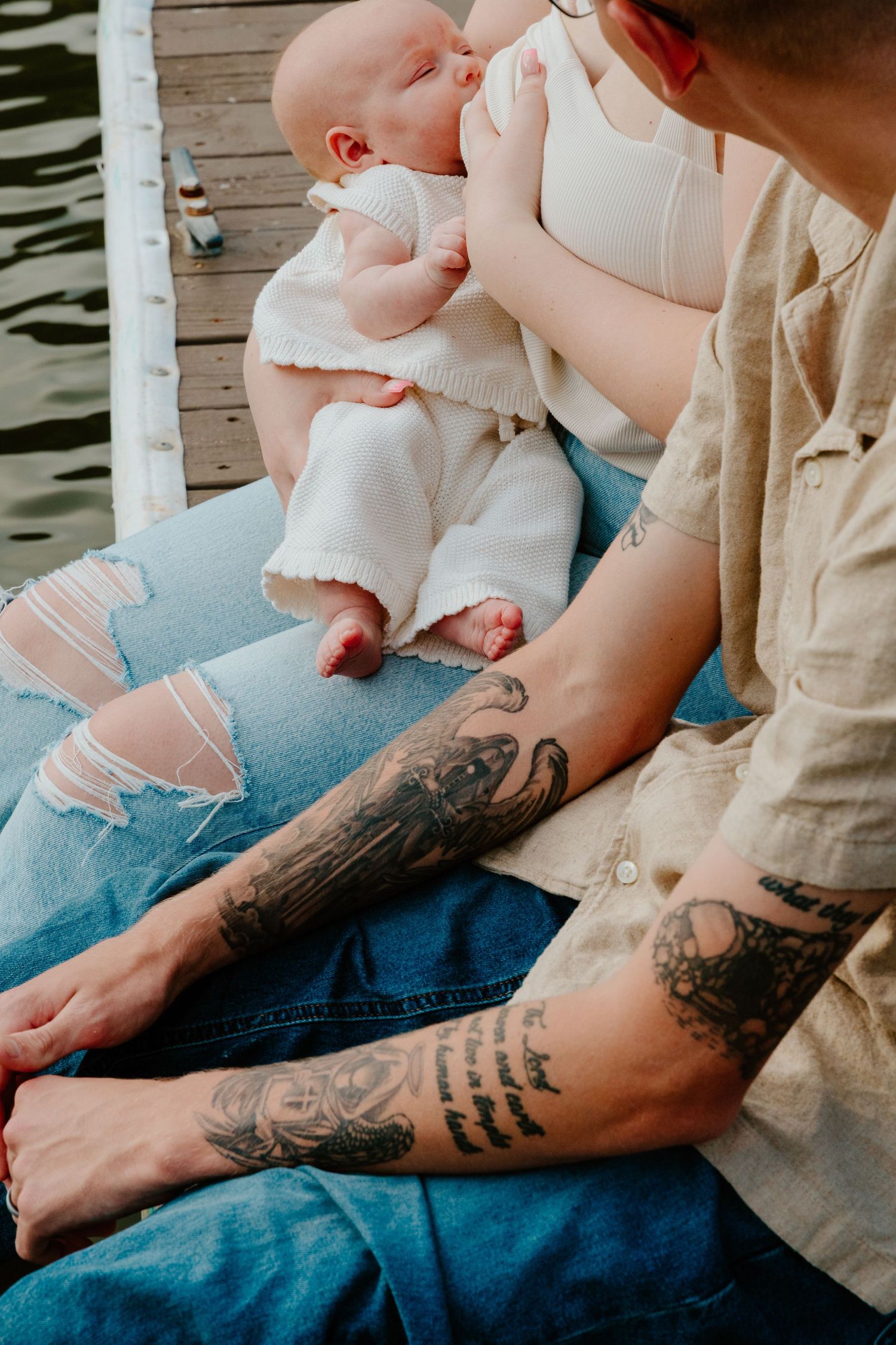 Close-up of tattooed parents sitting on dock holding their sleeping newborn baby.
