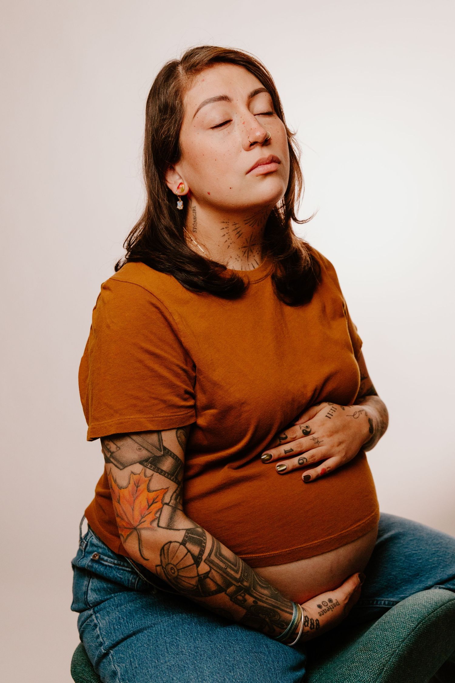 Tattooed pregnant woman in rust t-shirt holding belly with eyes closed peacefully.