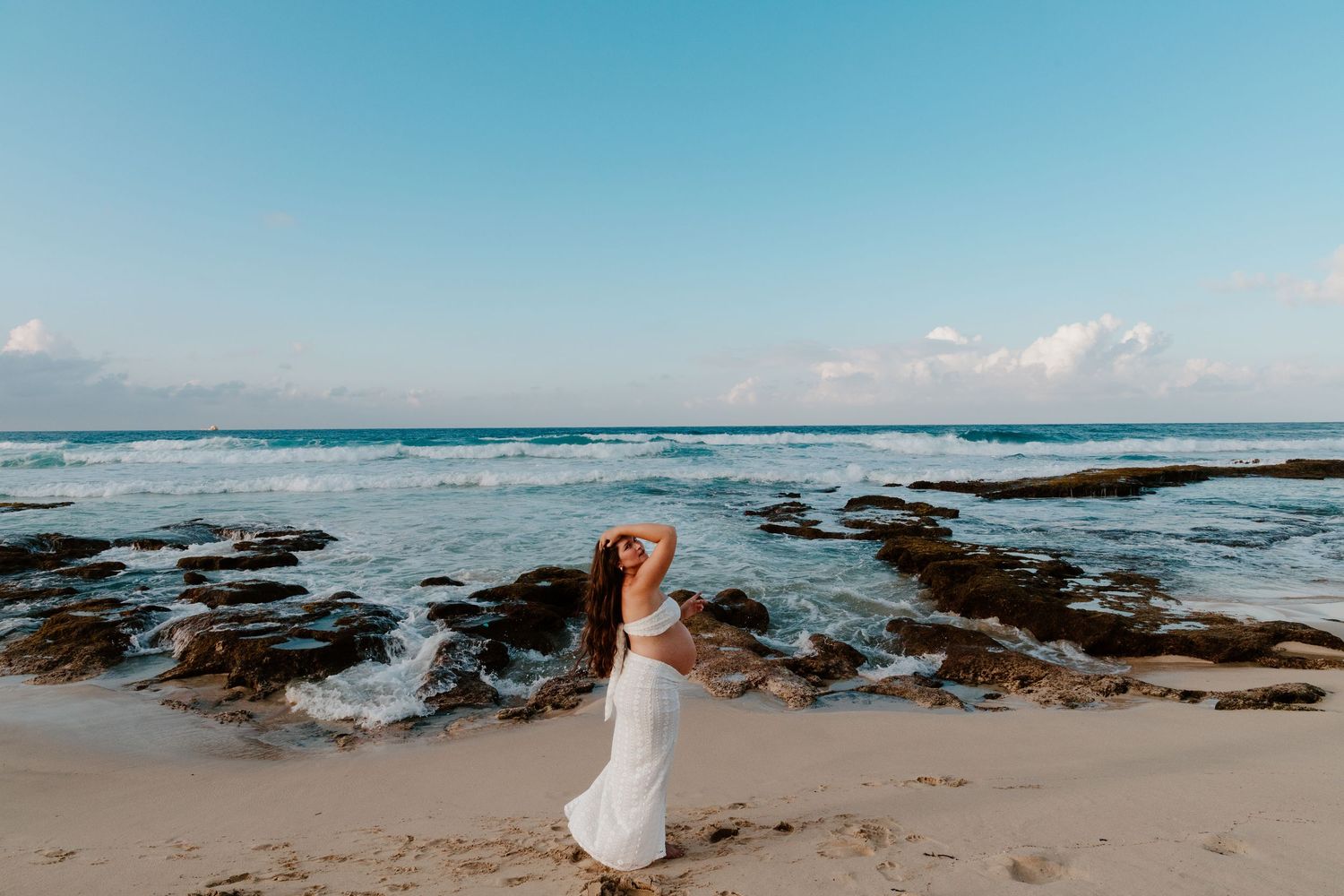 Pregnant woman in white outfit standing facing ocean waves at sunset beach.