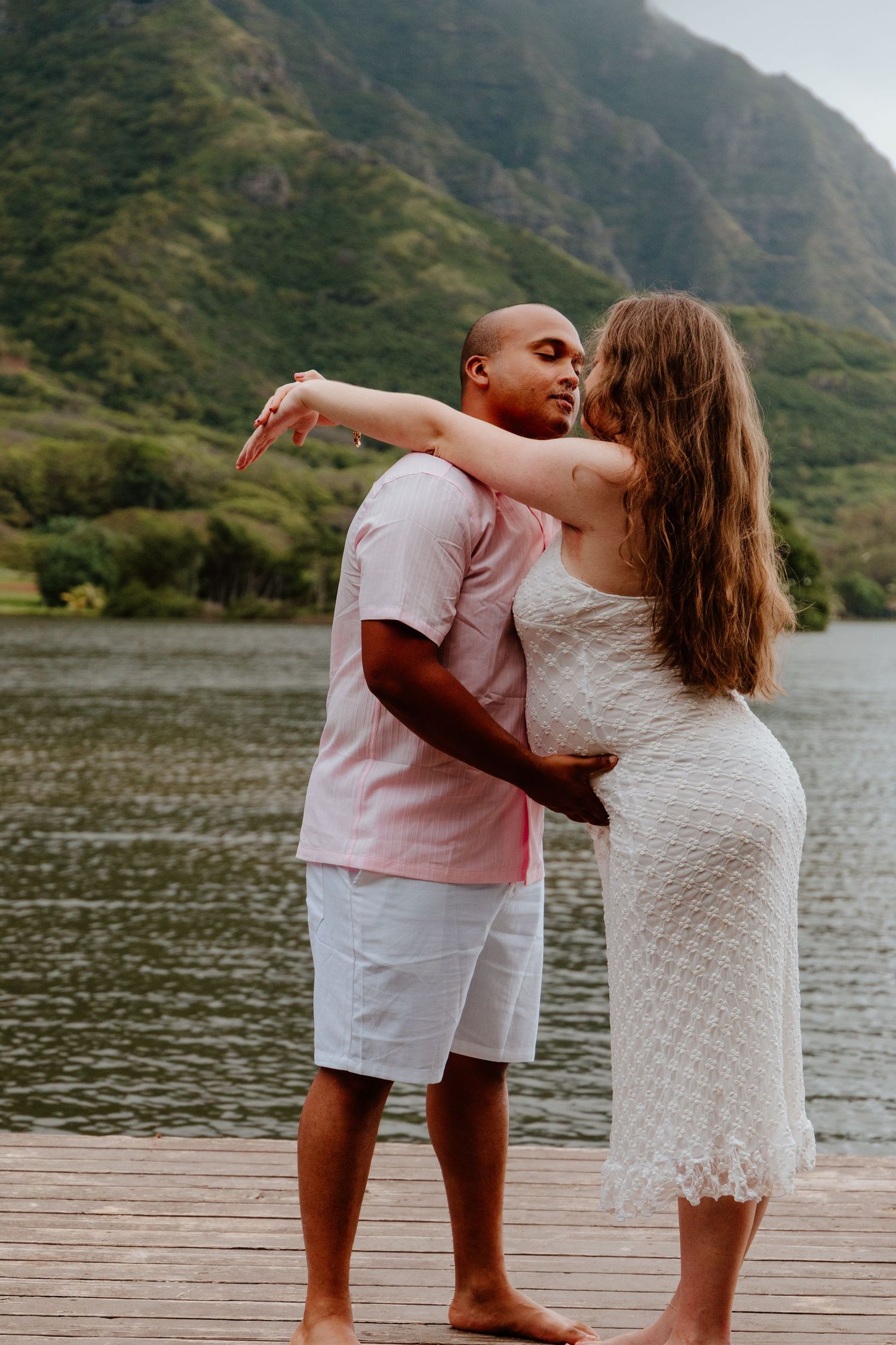 Romantic couple embracing and kissing on a wooden pier with a stunning mountain lake backdrop.