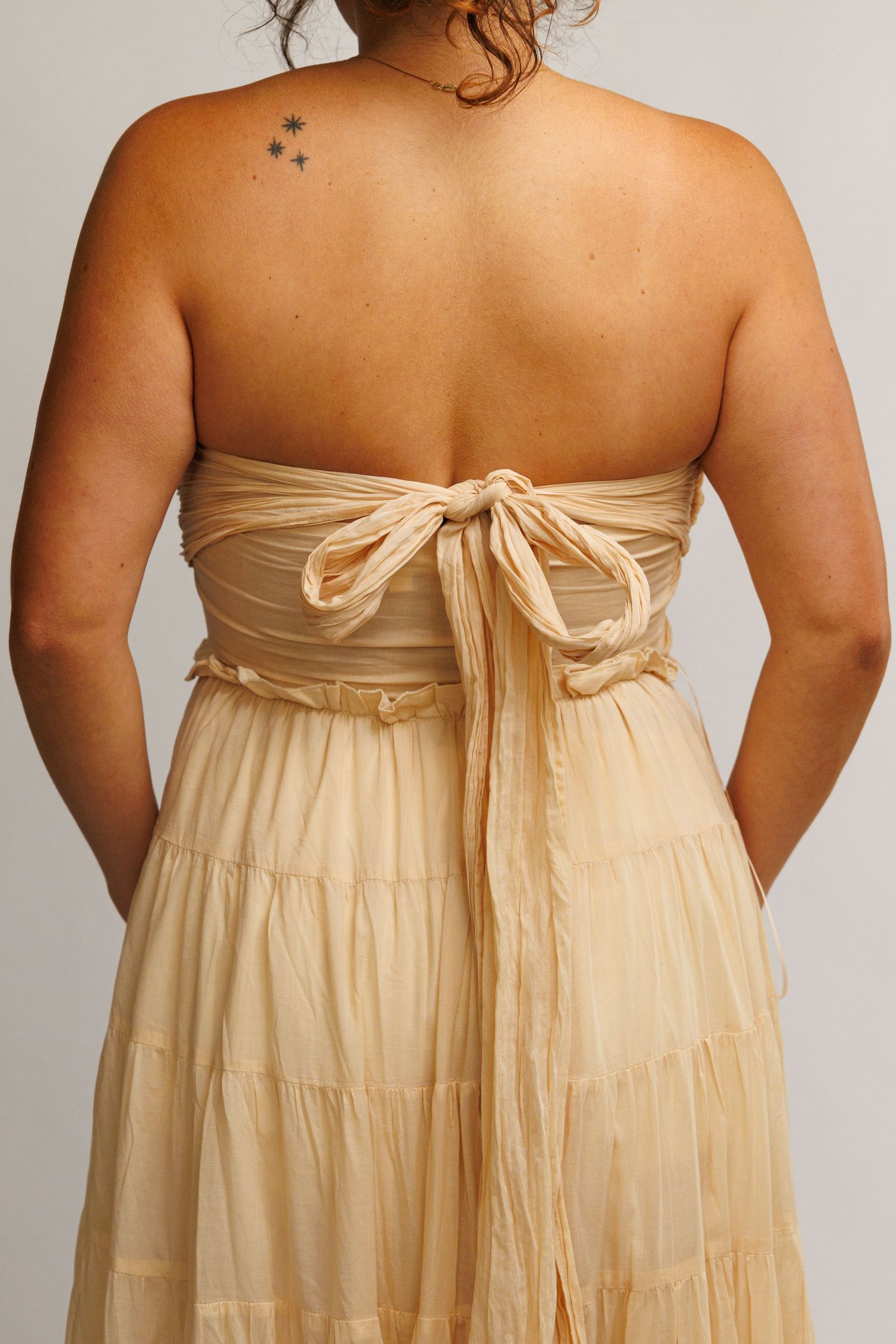 Back view of a woman in a strapless beige tiered maxi dress with a bow tie closure.