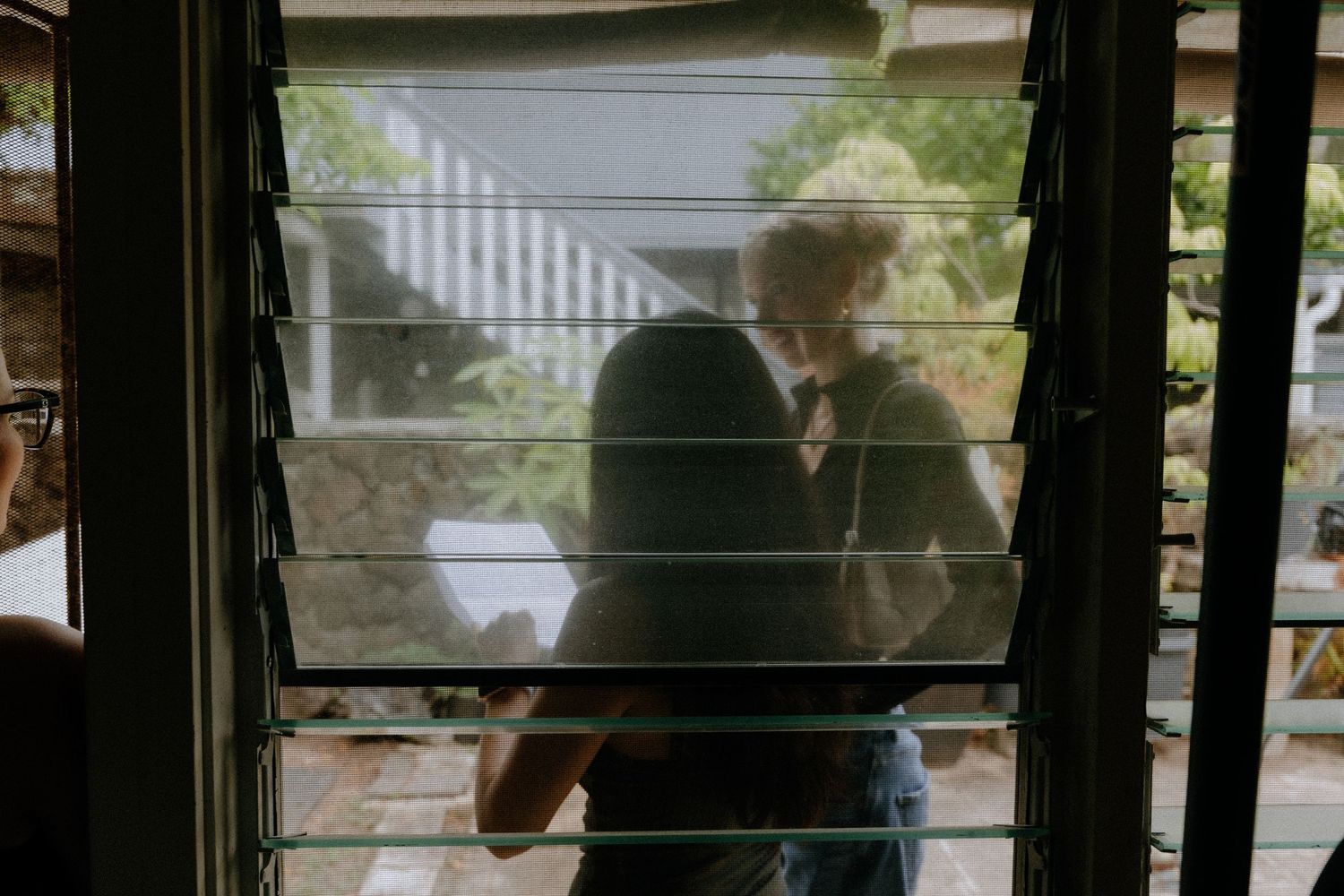 Silhouette of couple kissing behind window blinds with lush greenery visible outside.