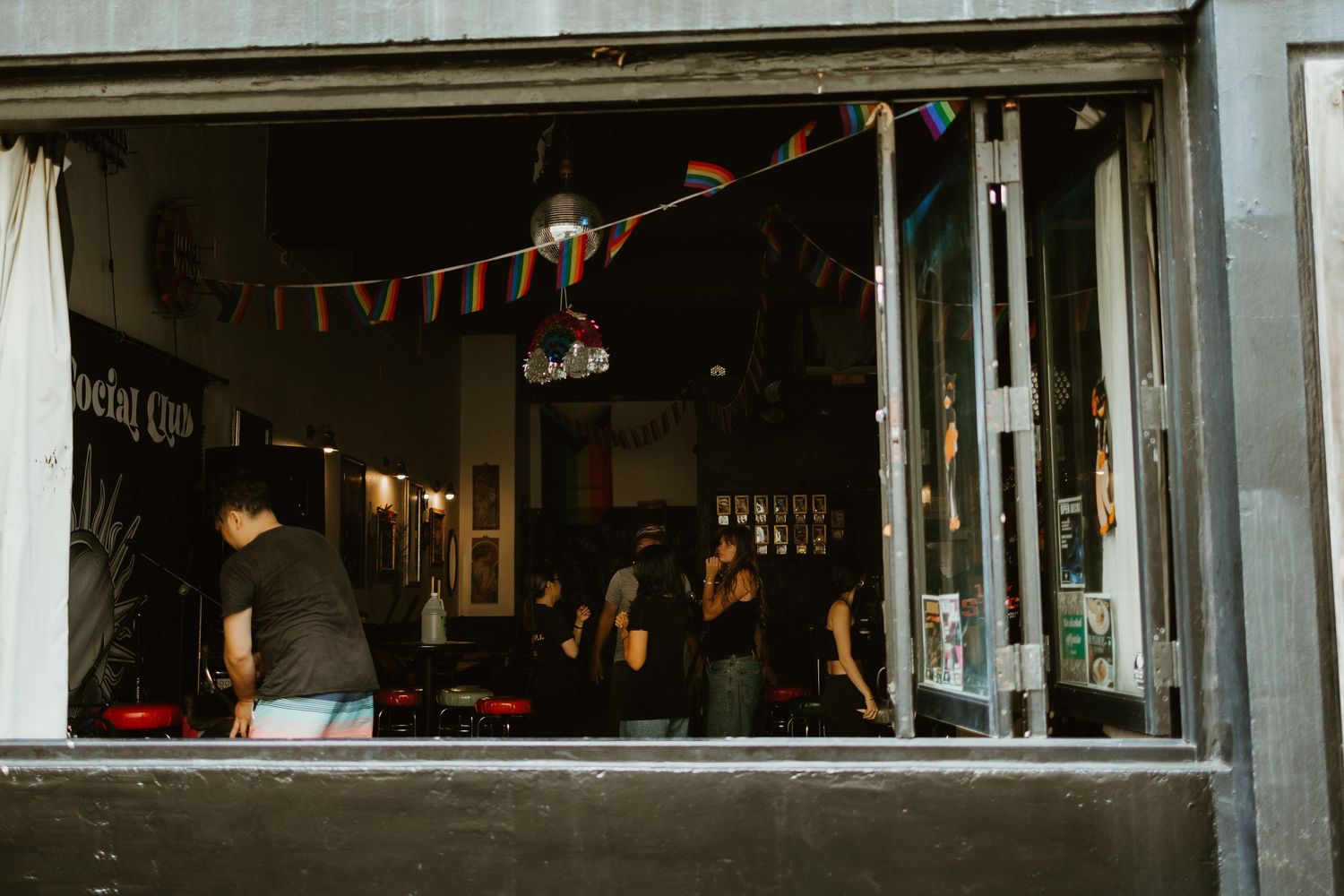 People socializing inside a dimly lit bar decorated with colorful bunting flags visible through a large window.