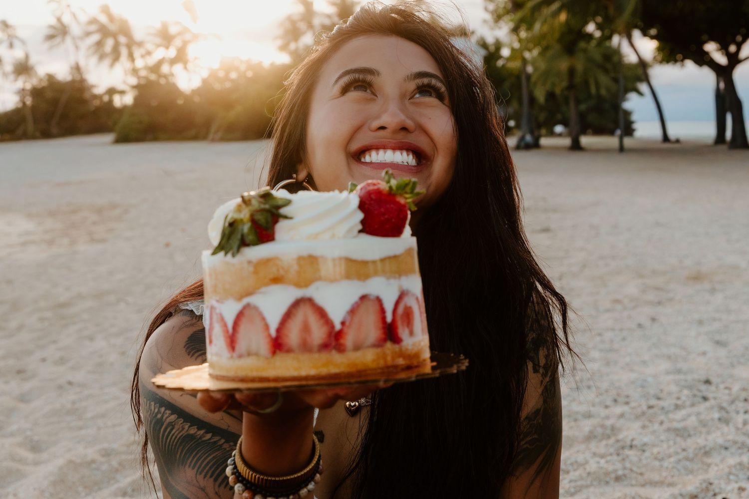 Smiling woman holding a beautiful strawberry layer cake on a sunny beach at golden hour.