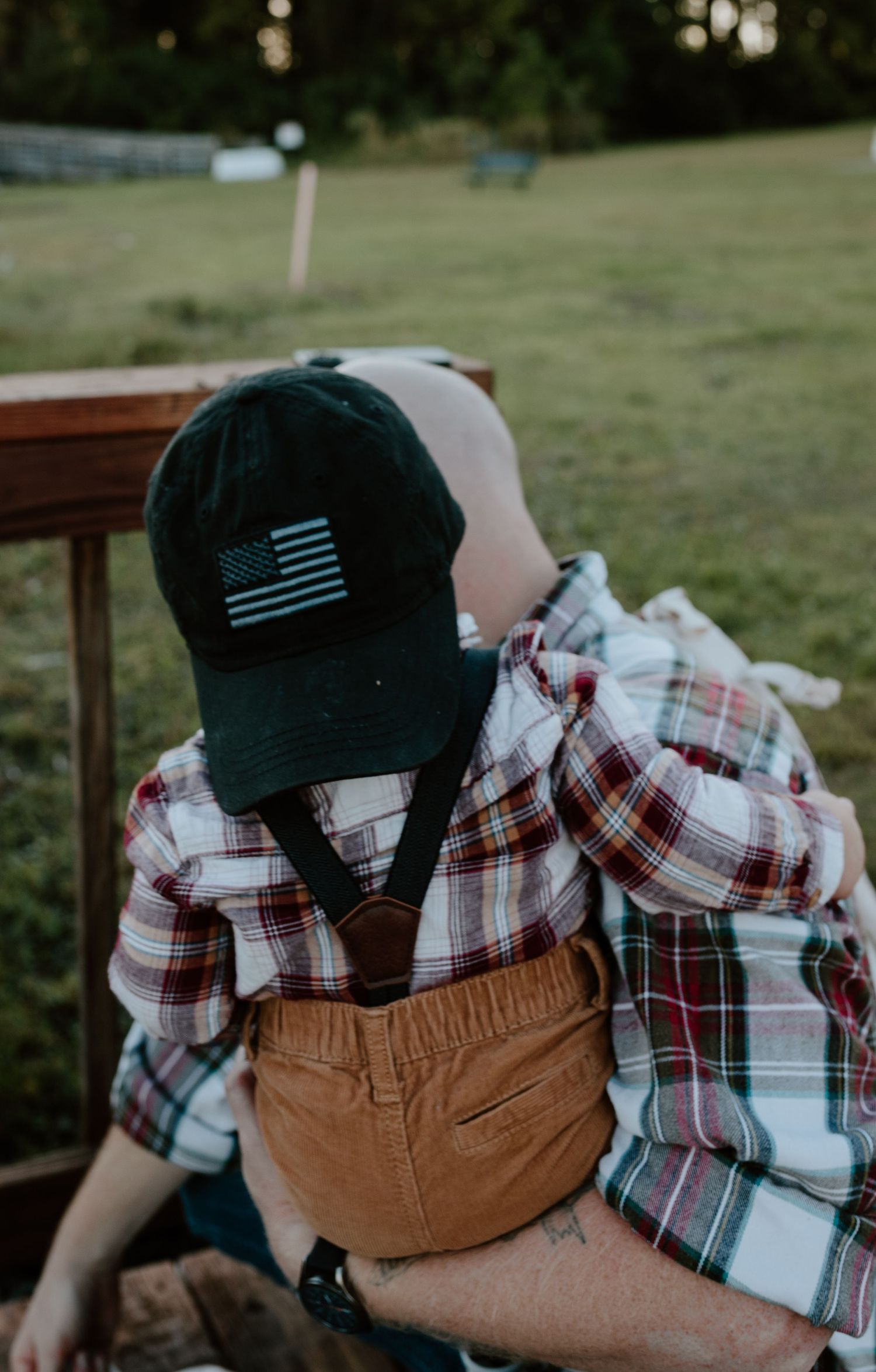 Young child wearing American flag cap and plaid flannel shirt with suspenders outdoors.