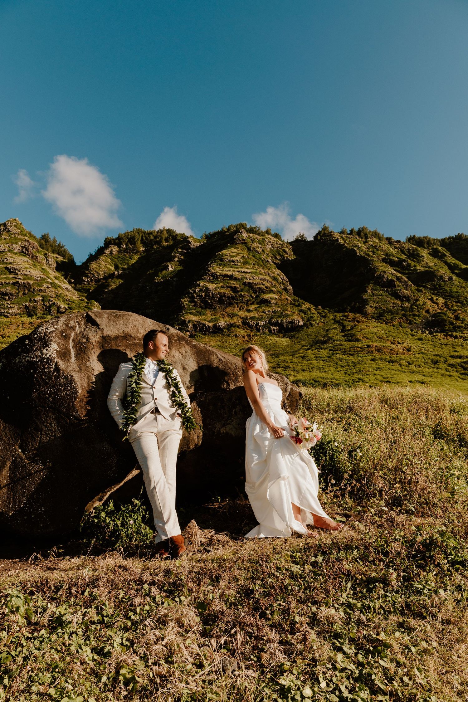 Bride and groom in white attire with floral leis pose against a large rock with lush green Hawaiian mountains behind.