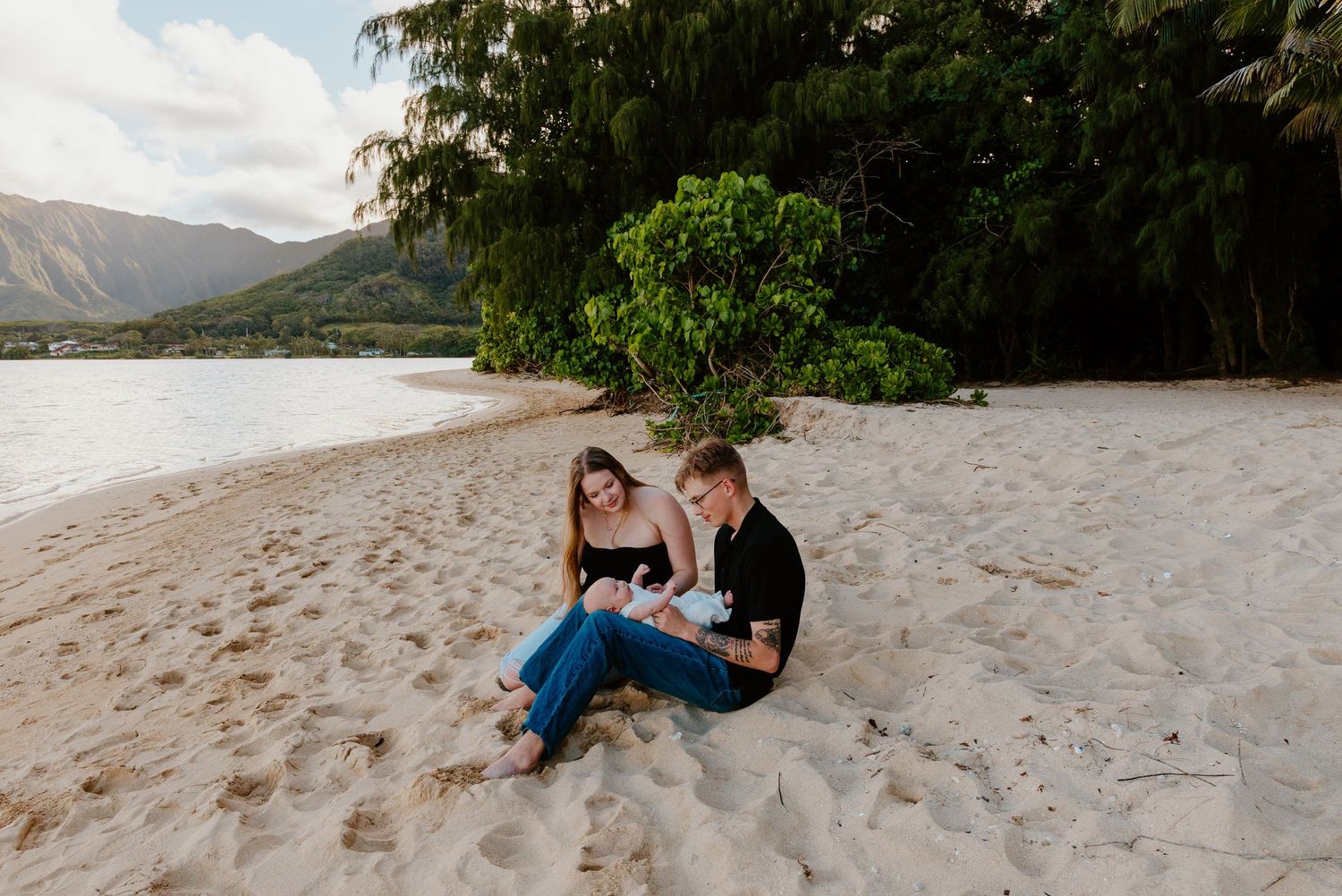 Young couple sitting on sandy beach with mountains backdrop cradling their newborn baby.
