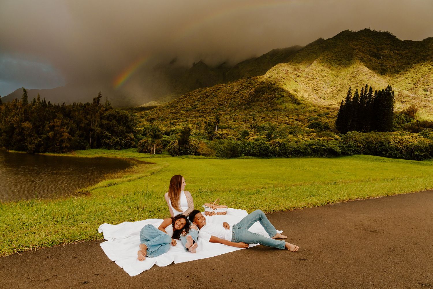Two people relaxing on a white blanket outdoors with a rainbow over misty mountains.