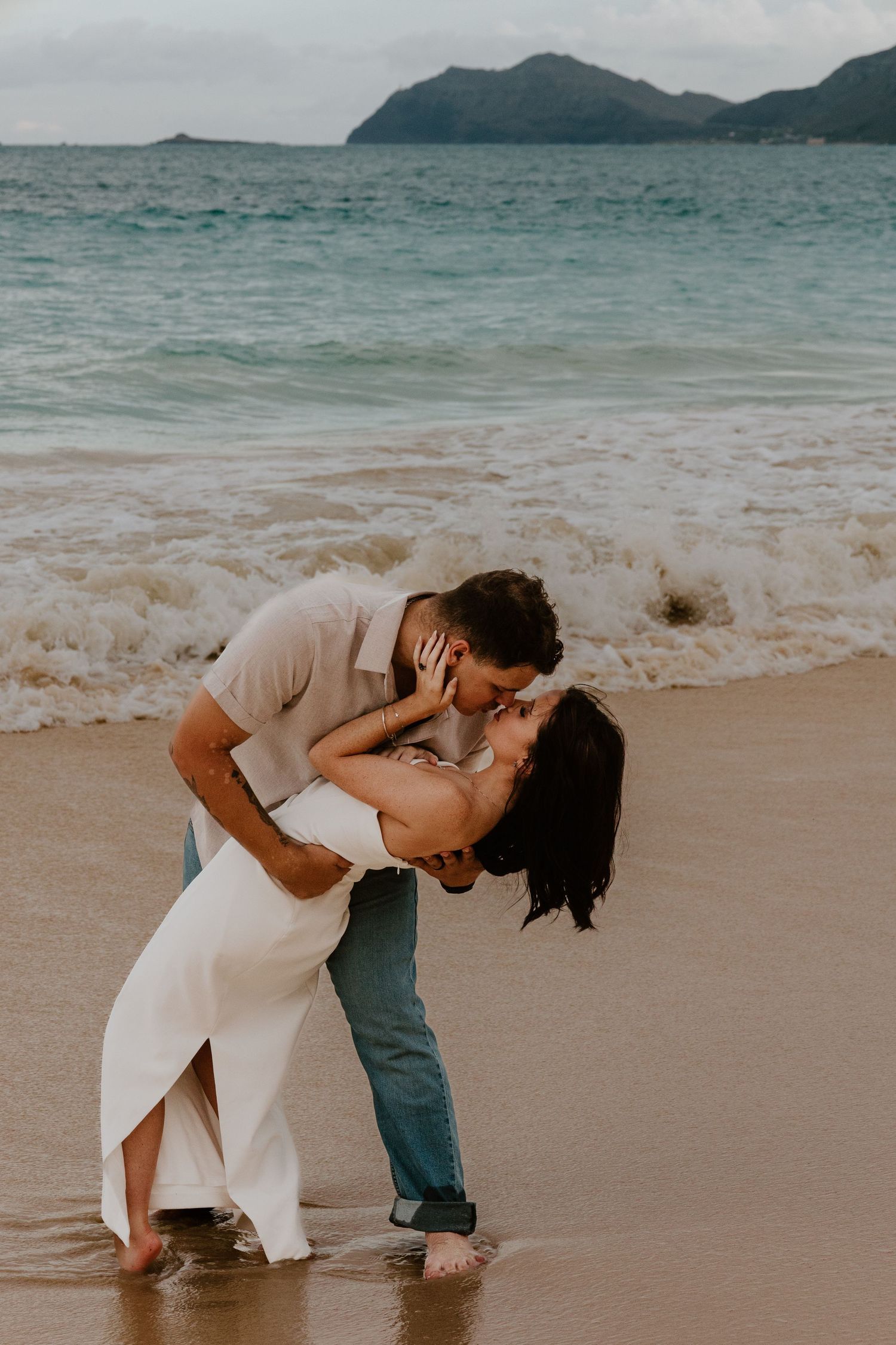 Couple sharing a romantic dip kiss on a Hawaiian beach with turquoise ocean waves and mountain islands in the background.