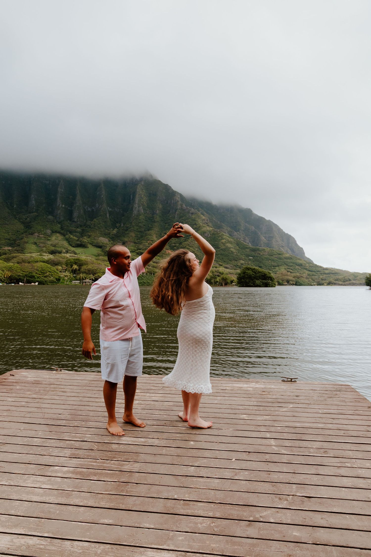 Couple dancing on a wooden dock with misty green mountains and a lake in the background in Hawaii.