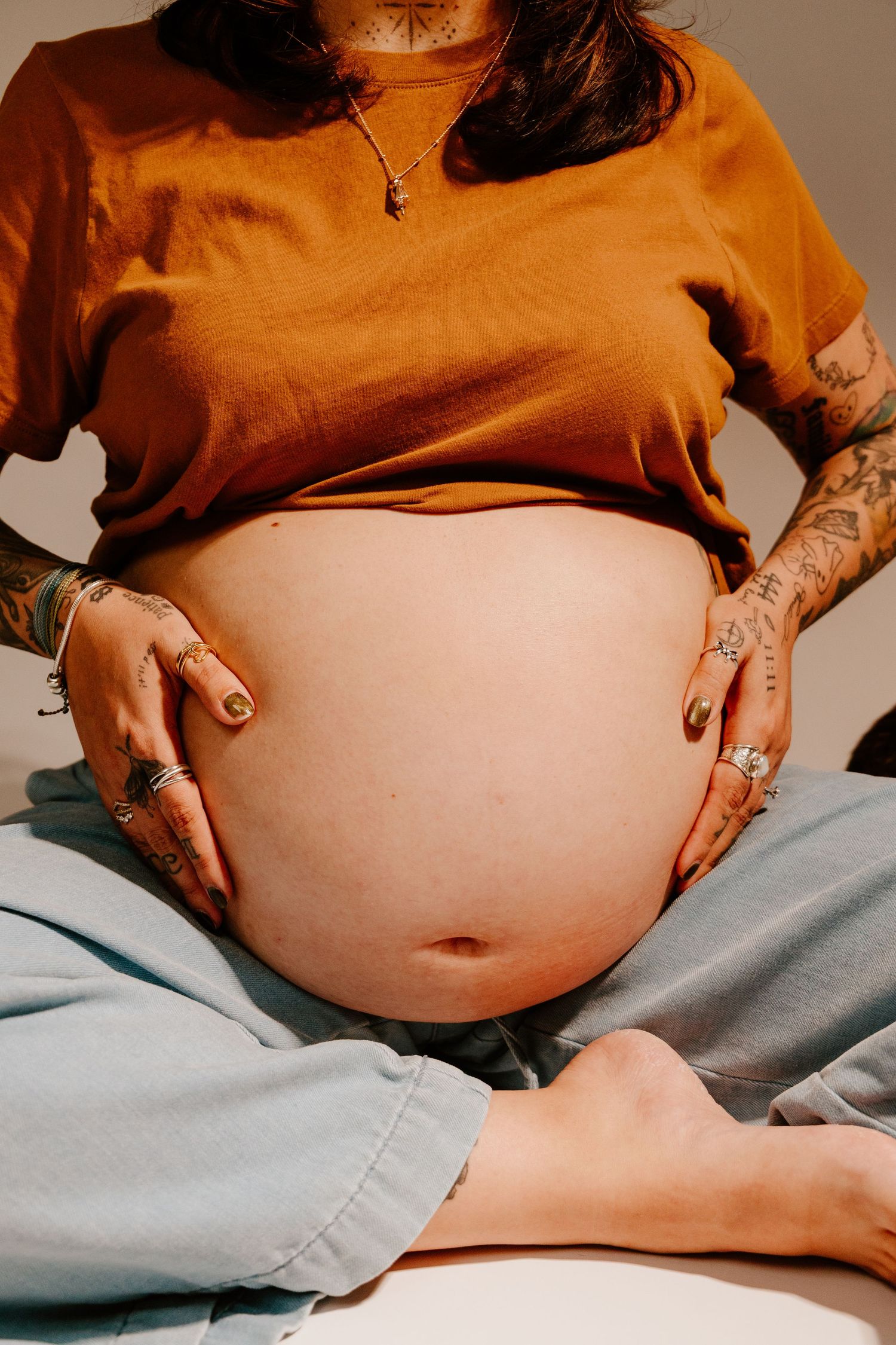 Pregnant woman with tattoos cradling bare baby bump, wearing orange shirt and jeans.