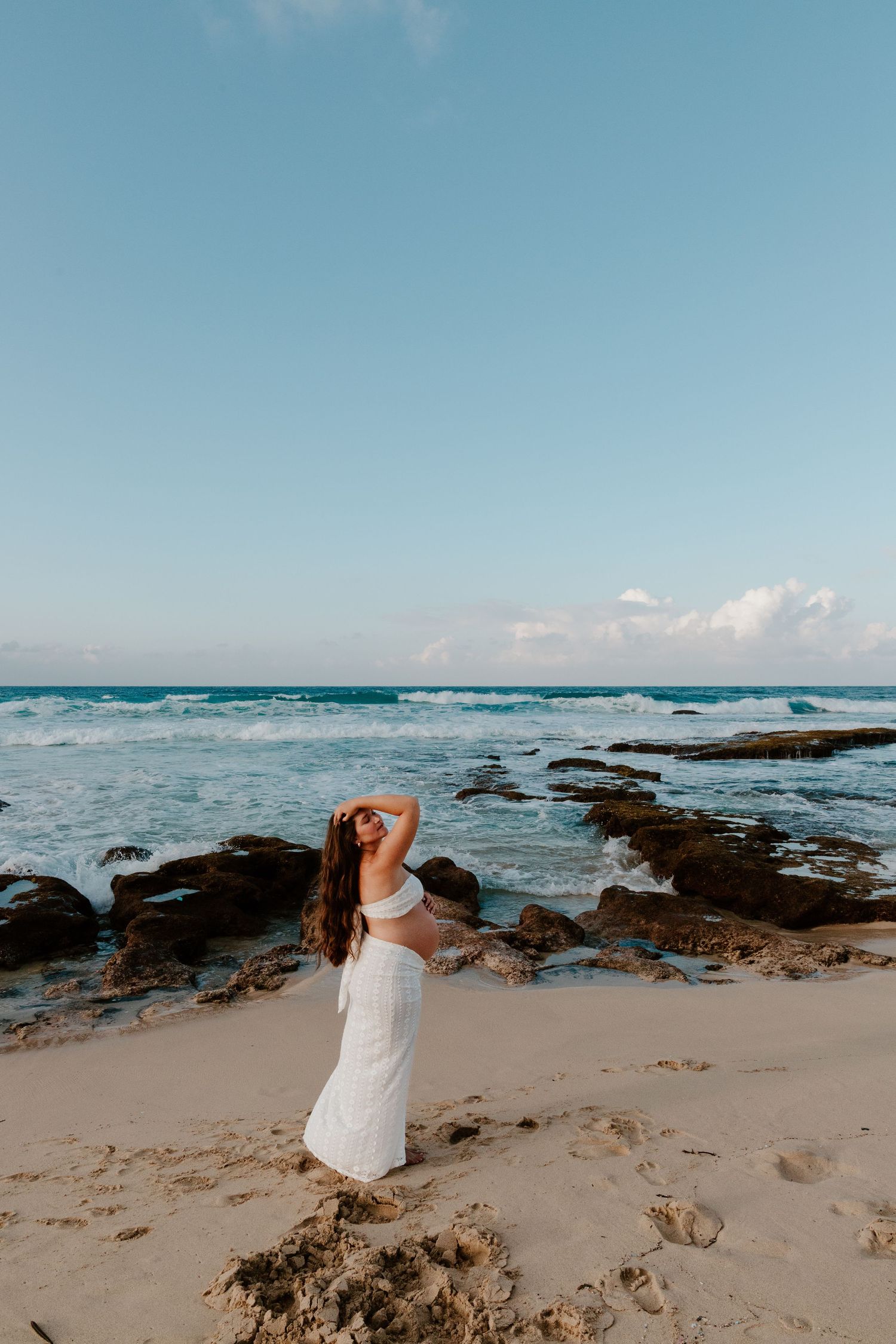 Pregnant woman in white crop top and skirt posing at rocky beach sunset.