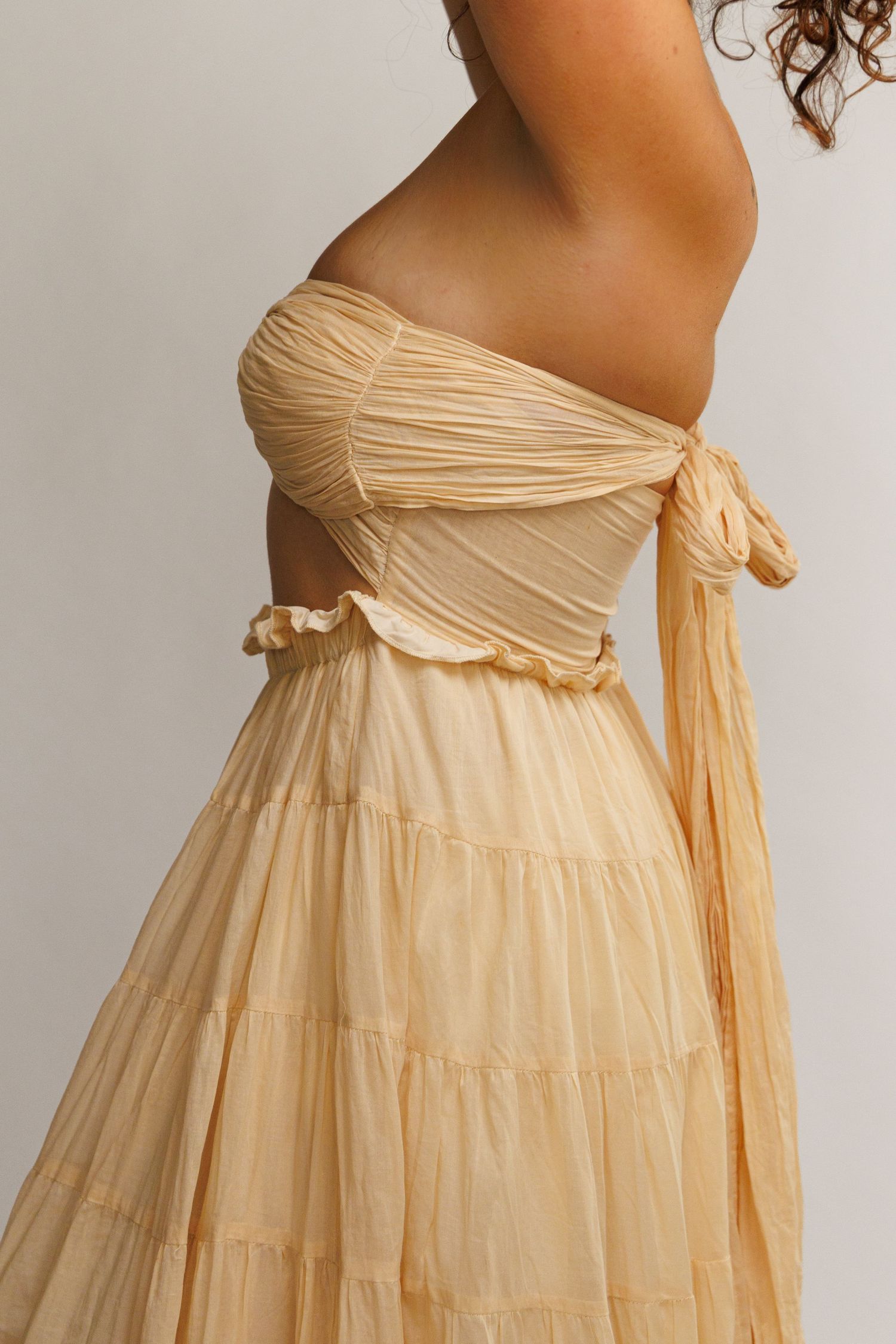 Woman wearing a flowing off-shoulder tiered maxi dress in soft champagne beige with ruched bodice and bow detail.