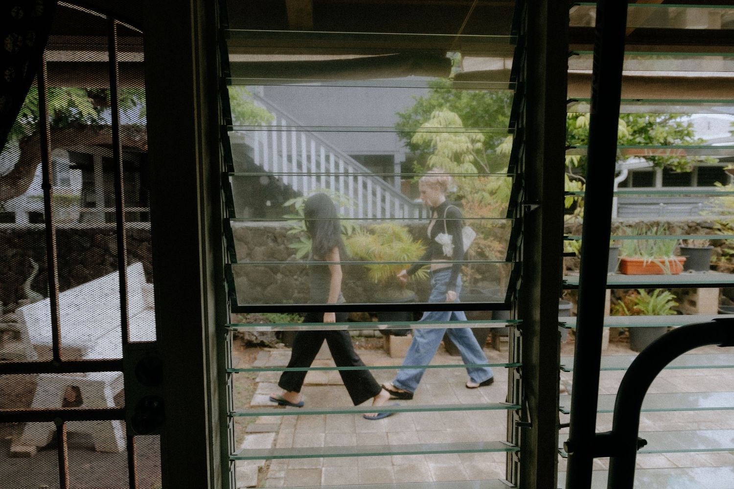 Two people walking past louvered glass doors with a tropical garden visible in the background.