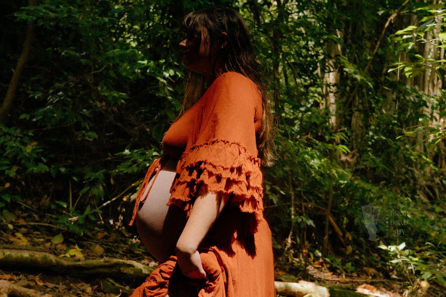 Pregnant woman in rust orange off-shoulder dress holding belly in lush forest setting.
