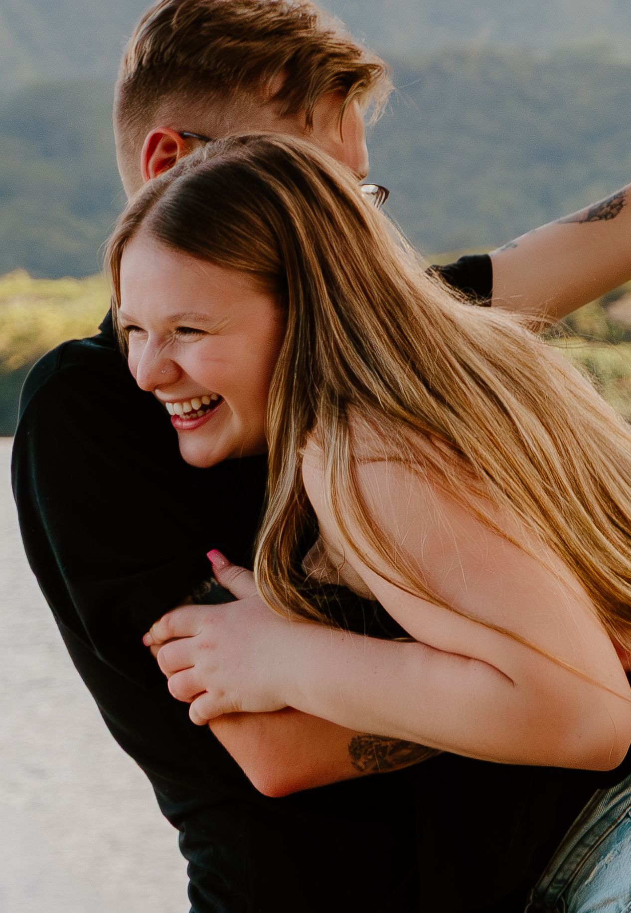 A joyful woman with long blonde hair laughs while embracing a man in a black shirt outdoors.