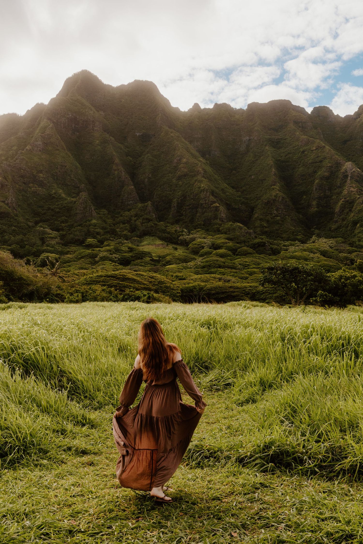 Woman in brown maxi dress walking through lush green field with dramatic Ko'olau mountain range backdrop in Hawaii.