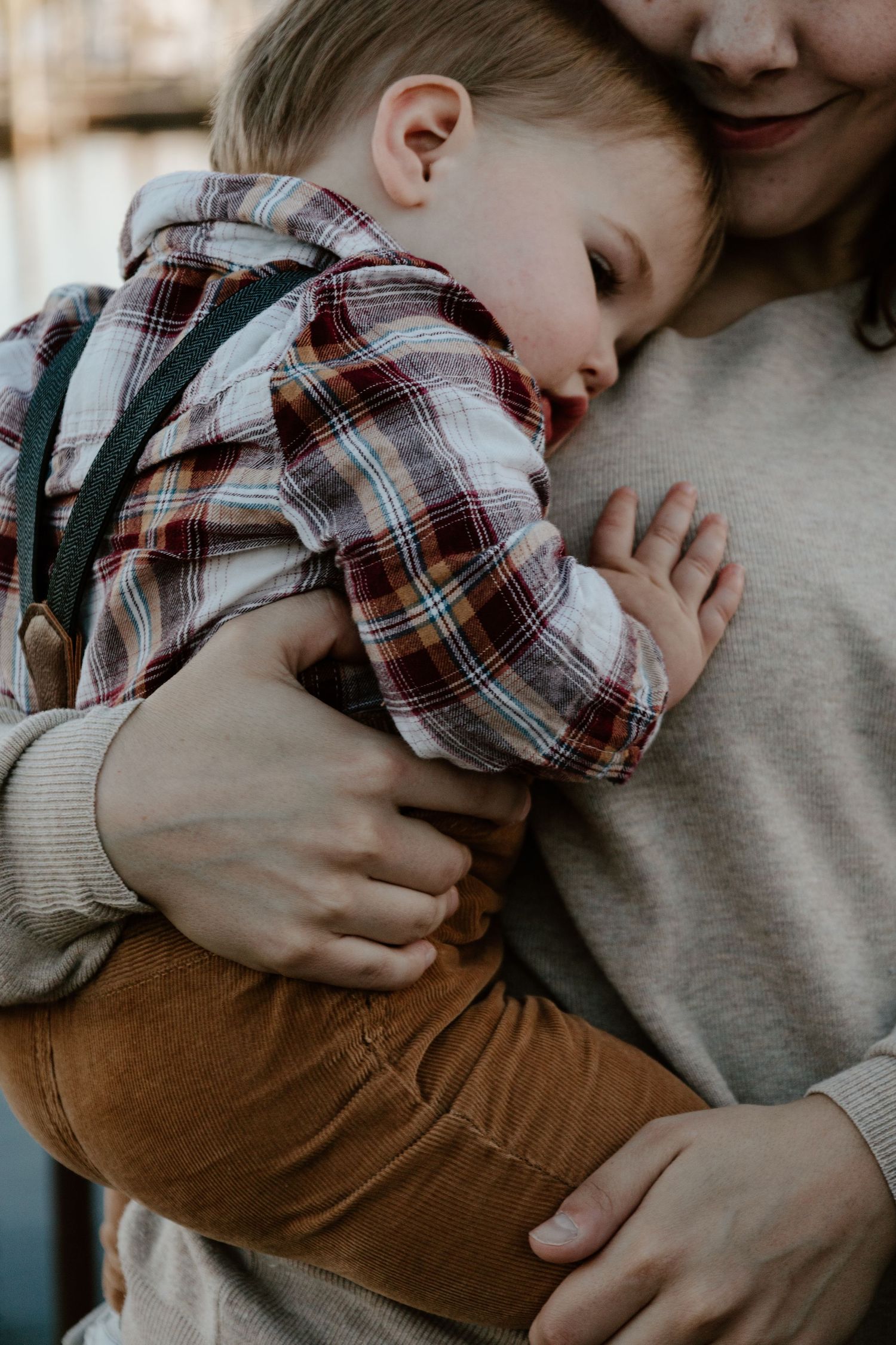 Mother in beige sweater gently holds sleeping toddler in plaid shirt against her chest in a tender embrace.