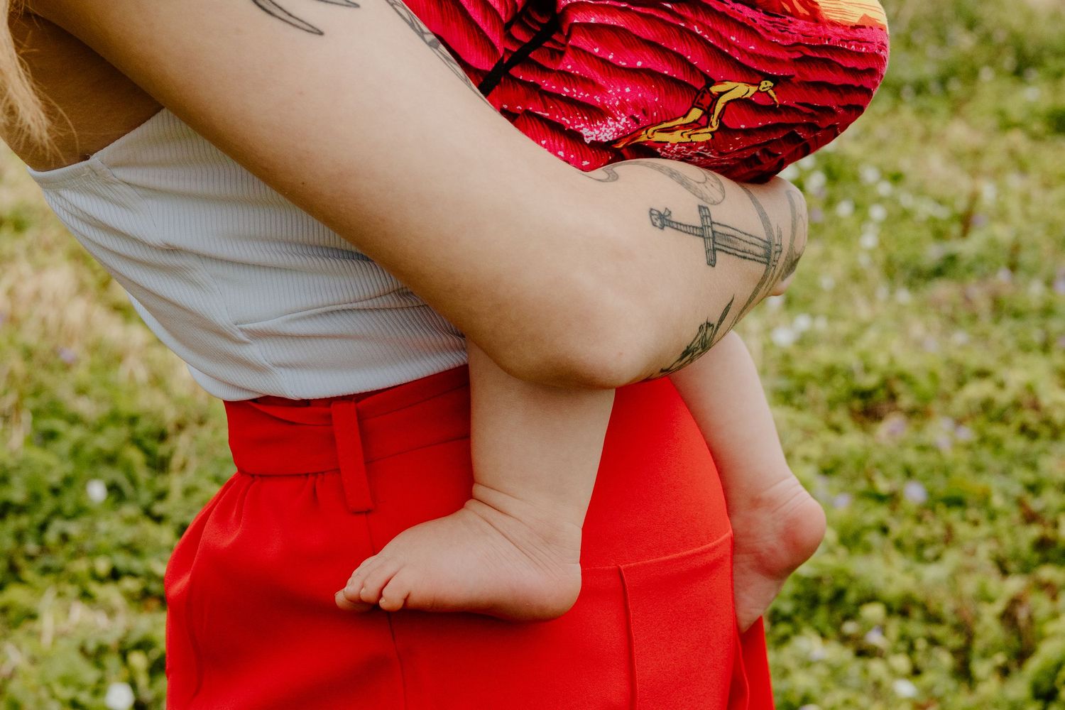 Tattooed parent holding baby in red dress with bare feet on green grass.