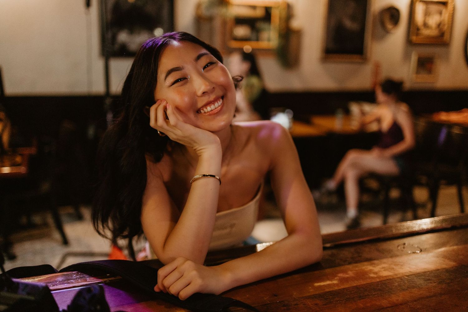 Smiling young woman with dark hair resting chin on hand at a wooden bar in a warmly lit restaurant.