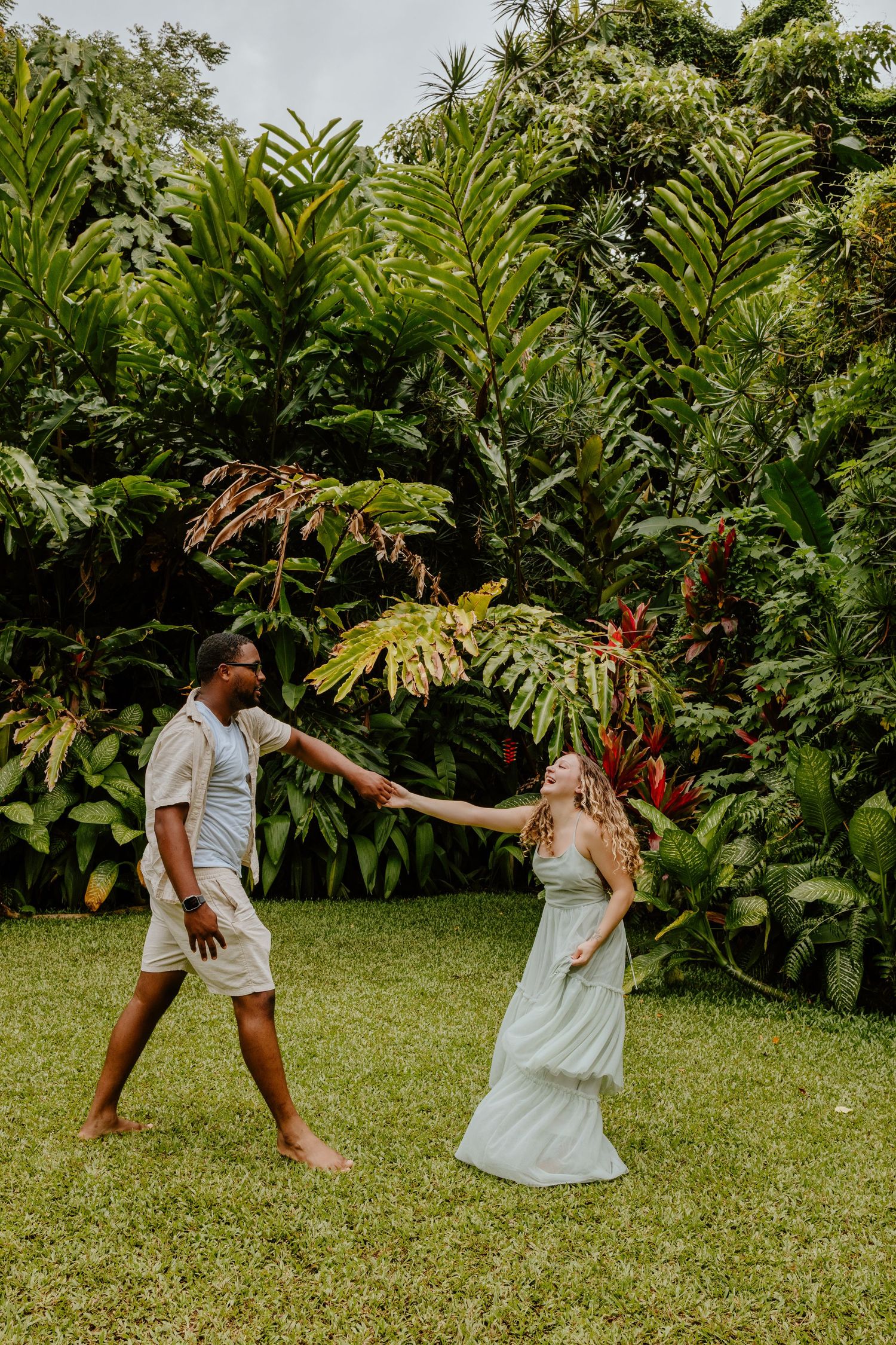 Couple dancing joyfully on green lawn surrounded by lush tropical foliage, wearing casual light-colored outfits.