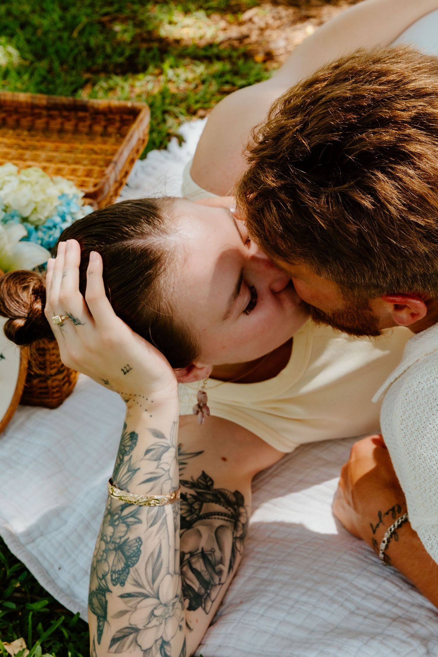 Couple sharing a romantic kiss during a picnic on a white blanket outdoors, woman with floral tattoos on her arm.