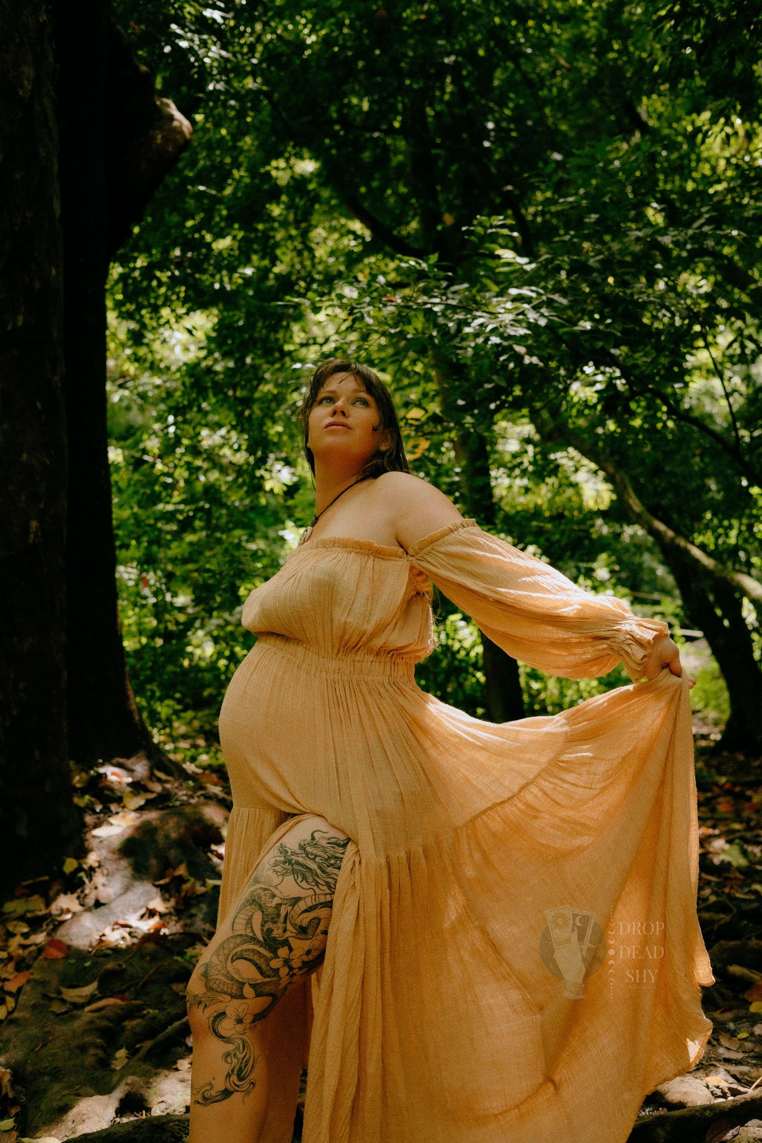 Pregnant woman in flowing peach dress holding skirt in lush green forest with tattoos visible.