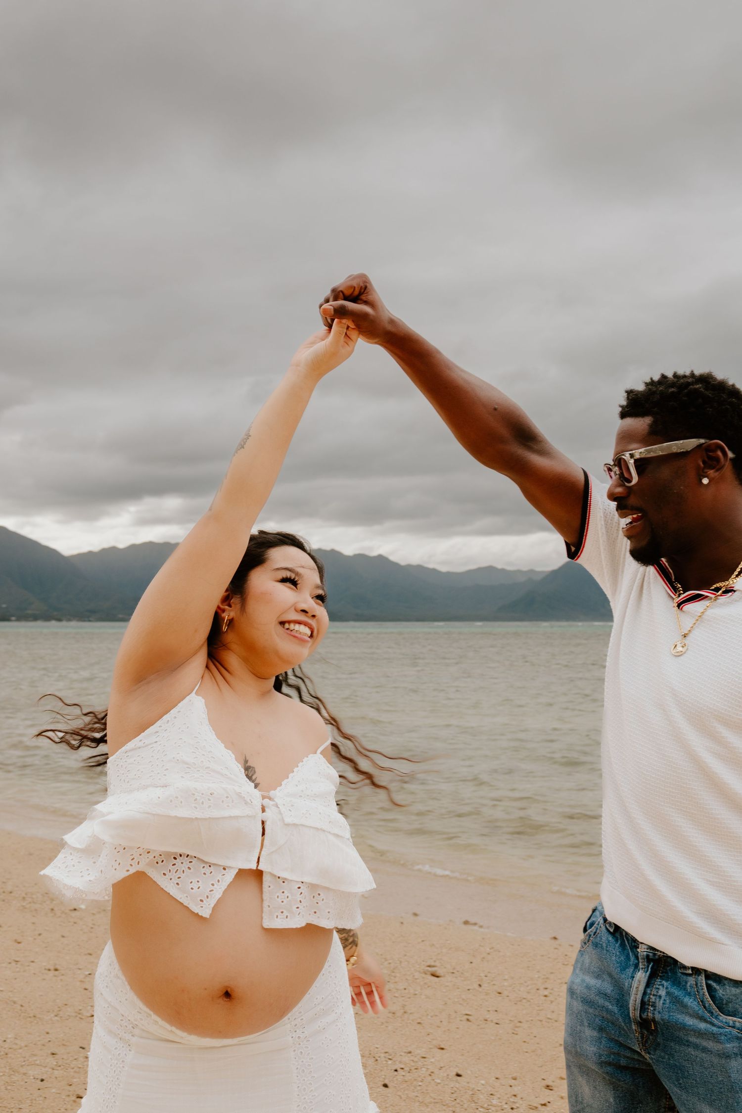 Interracial couple dancing on beach, pregnant woman in white crop top twirling.
