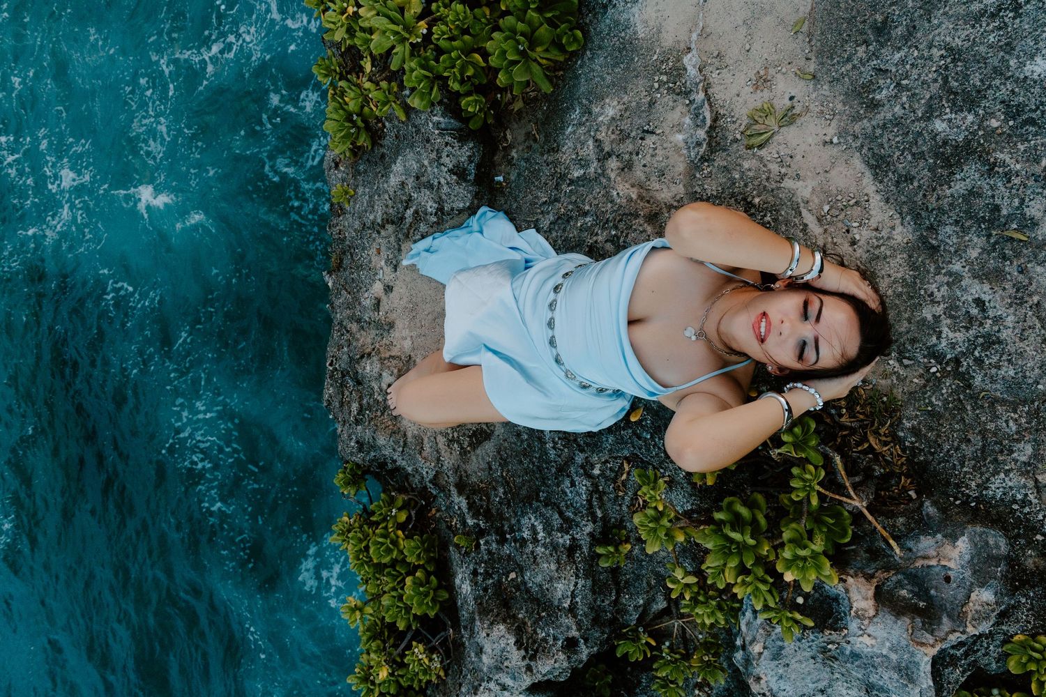 Aerial view of a woman in a light blue dress lying on rocky cliffs beside turquoise ocean waters.