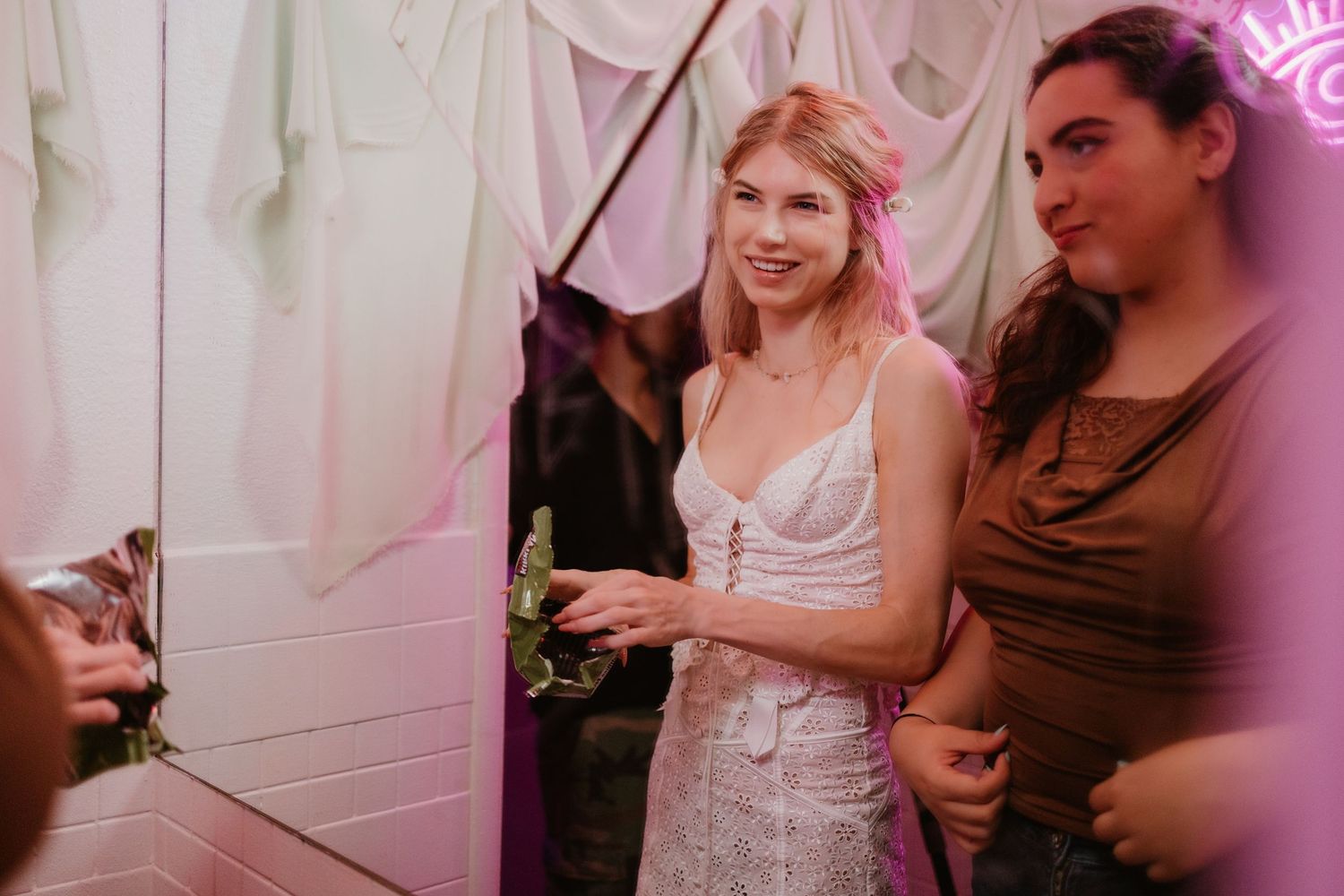 Woman in white lace dress smiling at a decorated indoor event with pink lighting and flowing fabric backdrop.