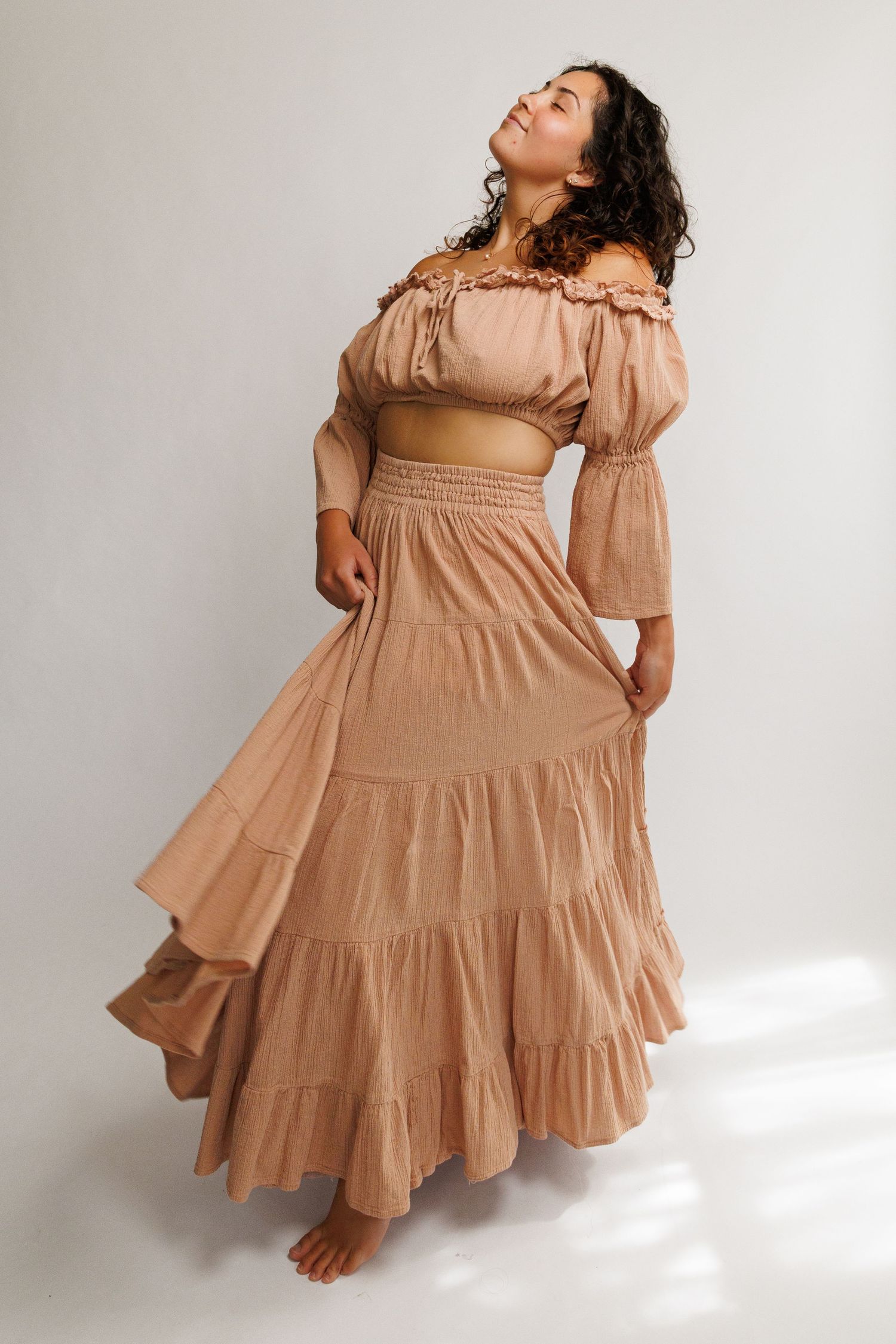 Woman wearing a tan off-shoulder crop top and tiered maxi skirt set, posing gracefully in a studio.