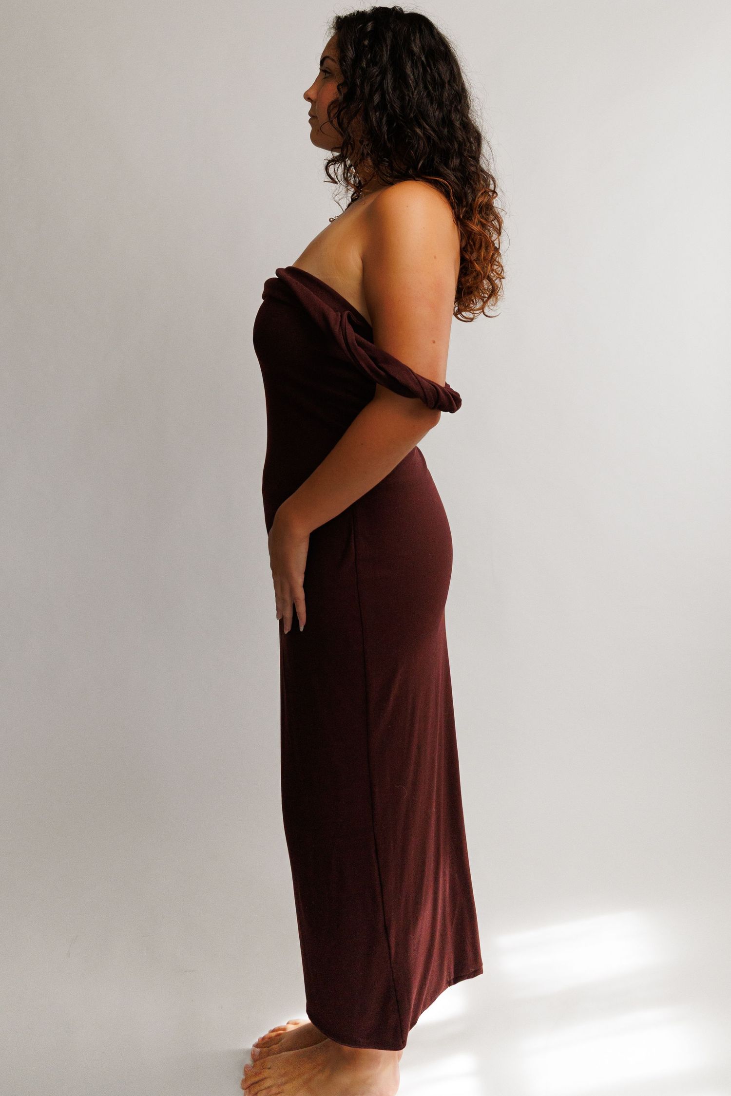 Woman wearing a dark brown off-shoulder maxi dress, form-fitting silhouette, side profile view.