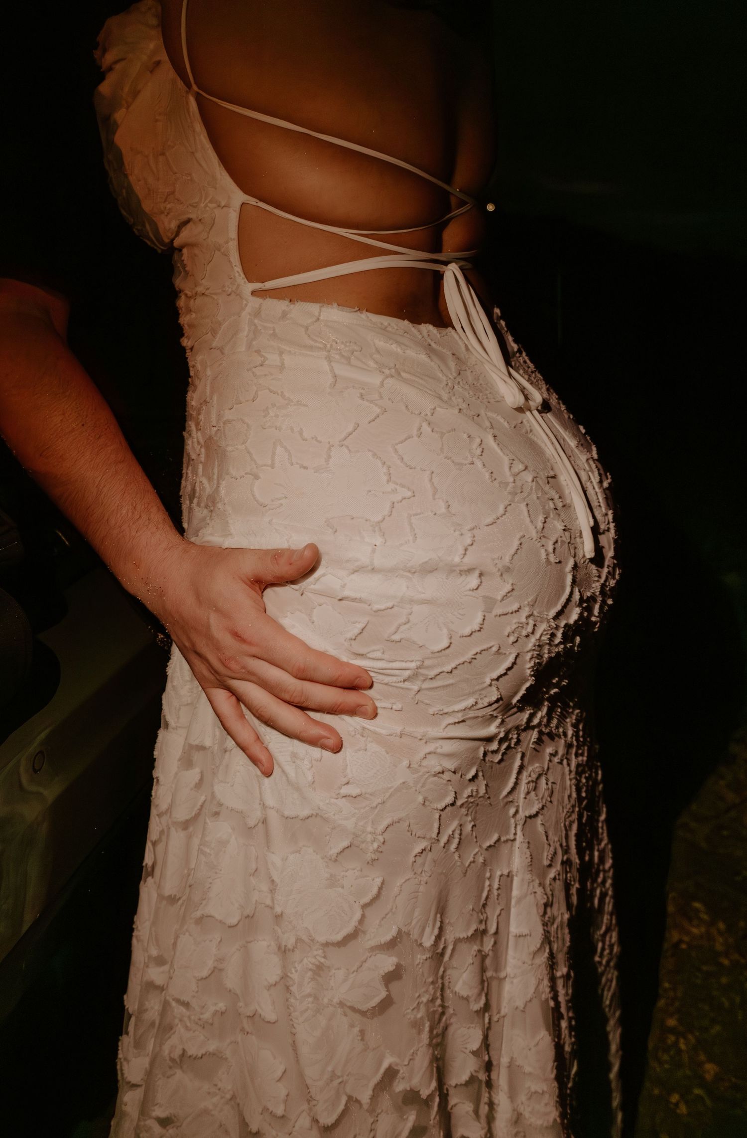 Close-up of a man's hand resting on a woman's hip in a textured white lace wedding dress.