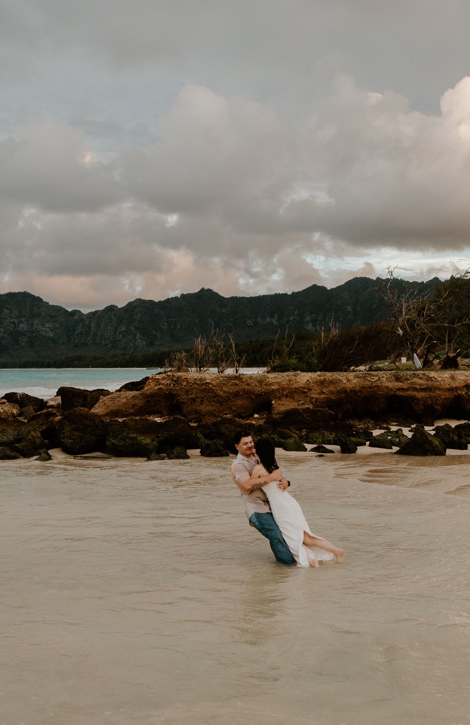 Man lifting woman in white dress while wading through shallow water with dramatic mountain backdrop in Hawaii.