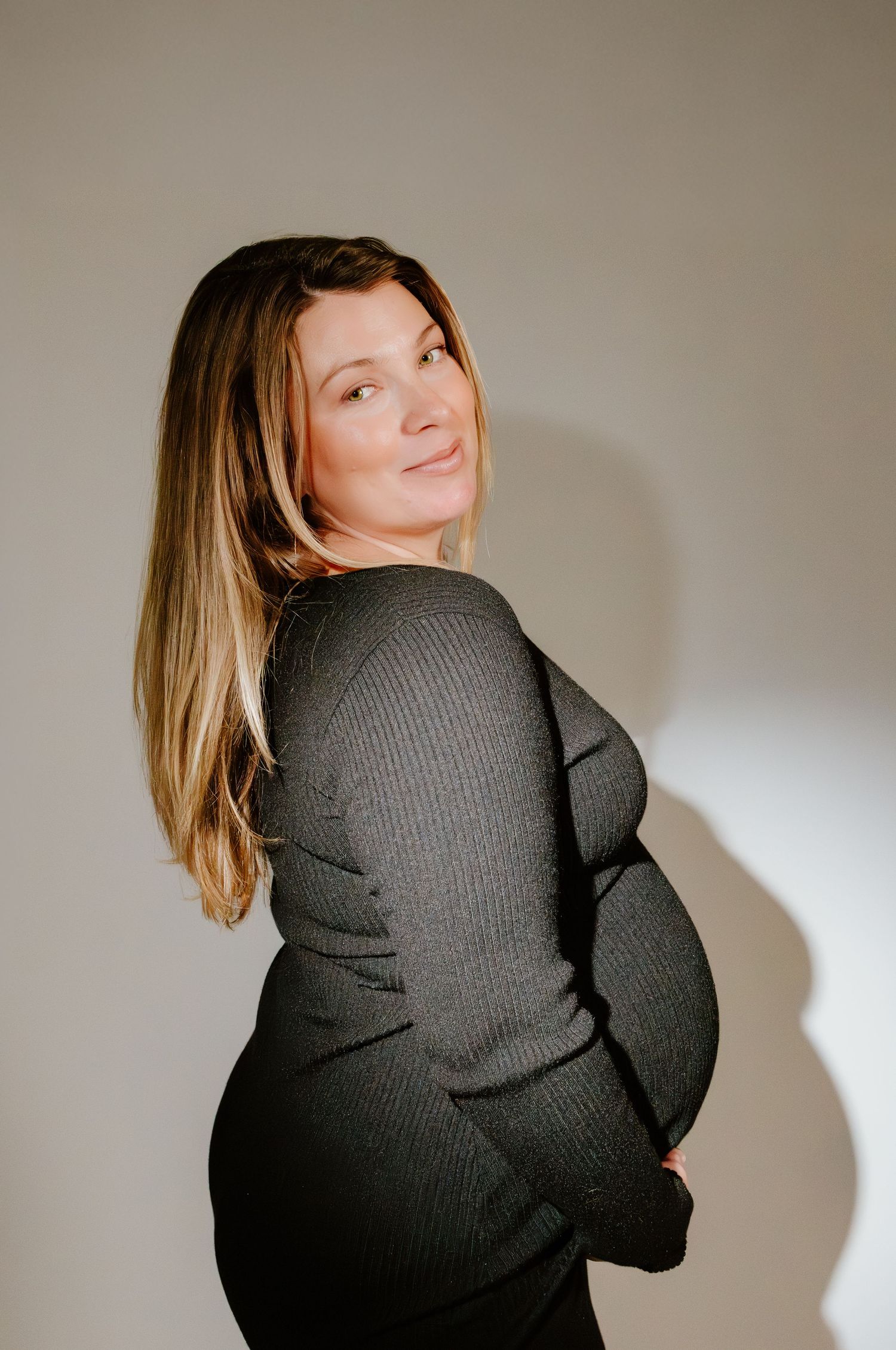 Smiling pregnant woman in dark ribbed dress cradling bump, glancing over shoulder in studio light.