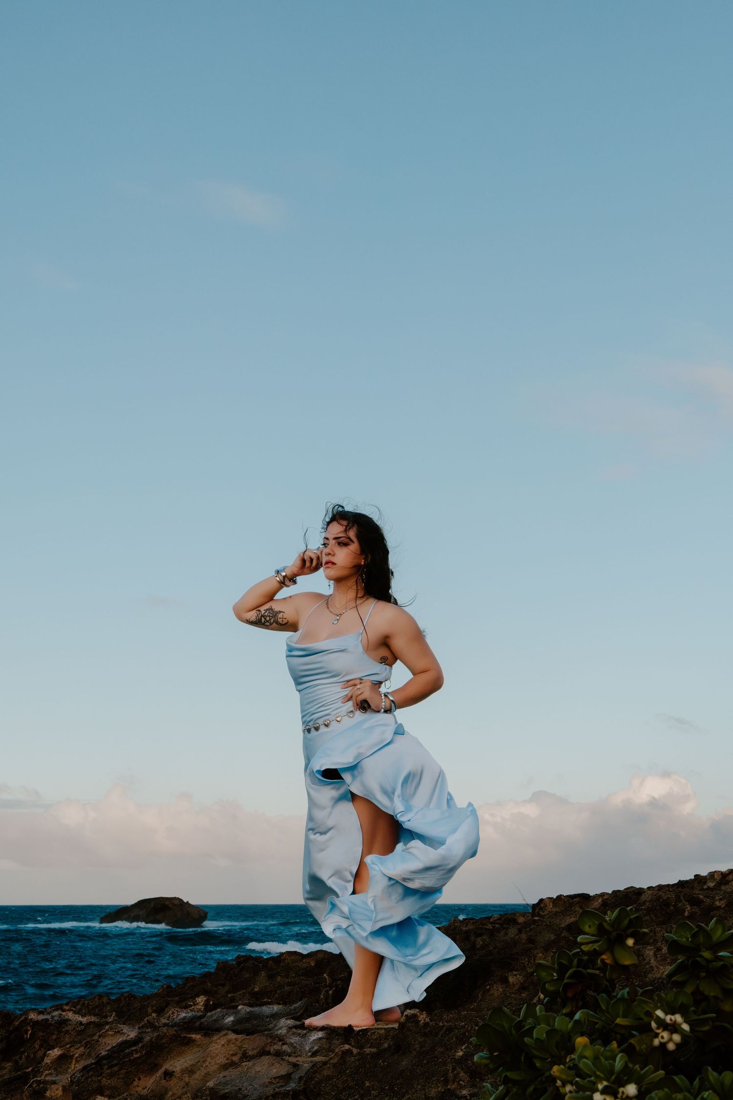 Woman in flowing light blue dress standing on rocky coastal cliffs with ocean and sky backdrop.
