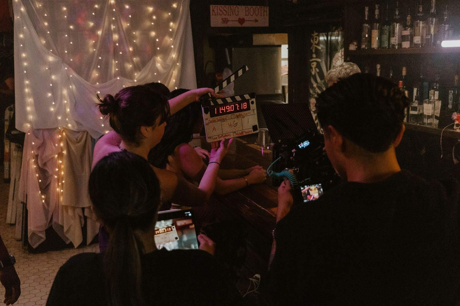 Film crew on a movie set holding a clapperboard in a dimly lit bar with string lights in the background.