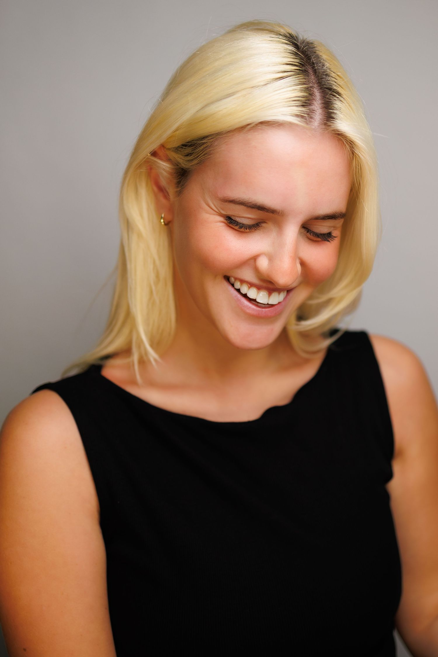 Smiling blonde woman with dark roots wearing a black sleeveless top against a gray background.