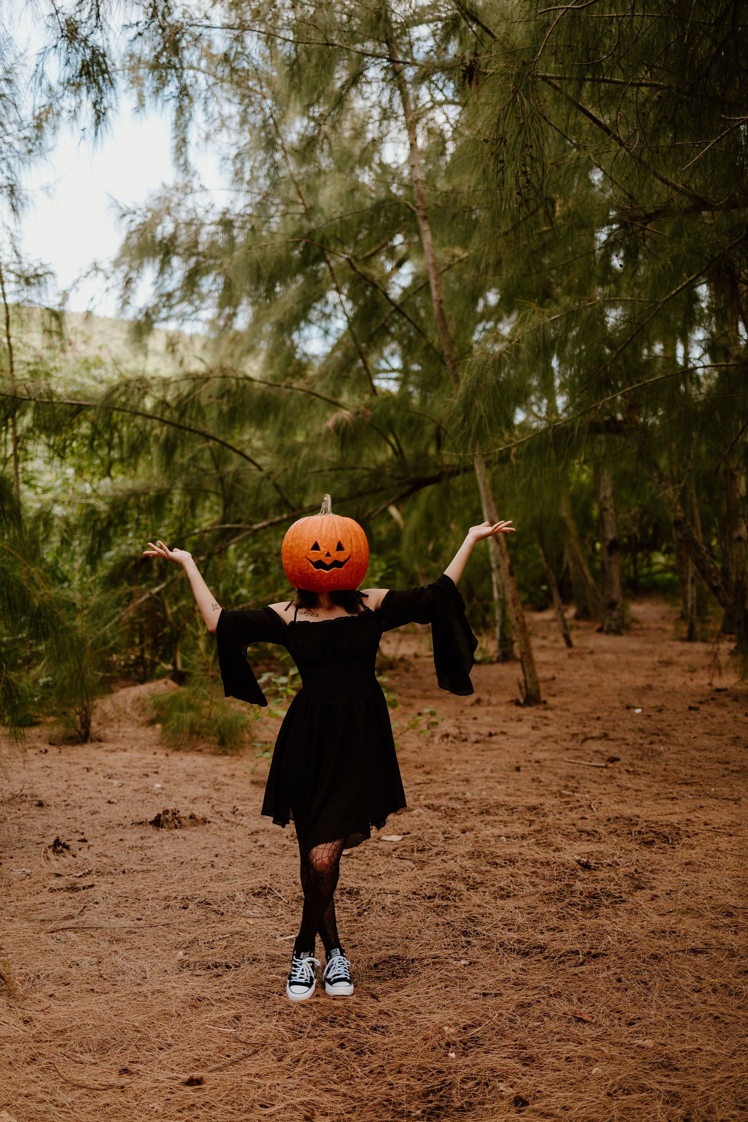 Person in a black dress and sneakers with a carved jack-o'-lantern head poses with arms raised in a sunlit pine forest.