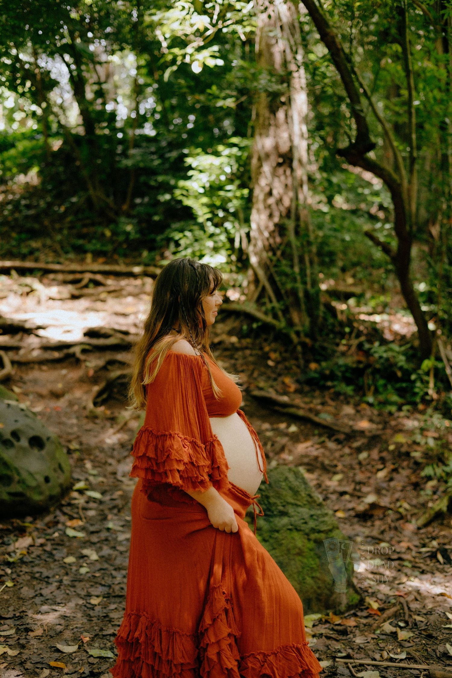 Pregnant woman in rust orange ruffled dress cradling belly on forest path among trees.
