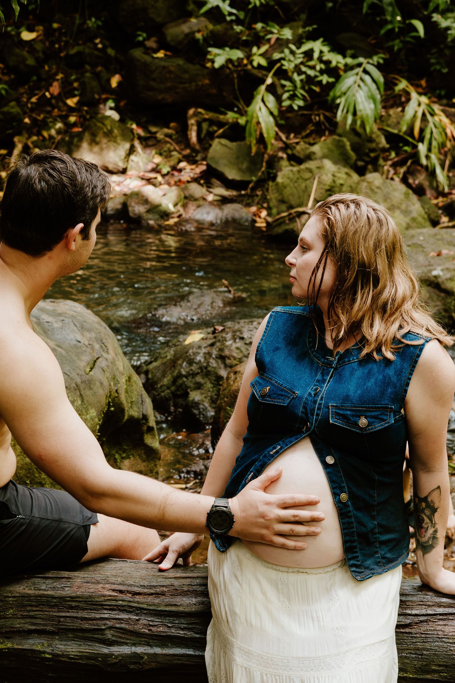 Couple sitting on log in forest creek, man touching pregnant womans exposed belly in denim vest.