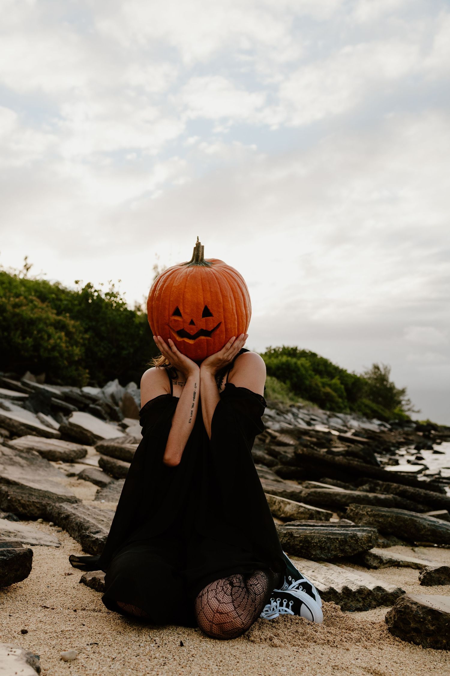 Person in a black dress and fishnet tights kneels on a rocky beach holding a carved jack-o'-lantern pumpkin over their face.