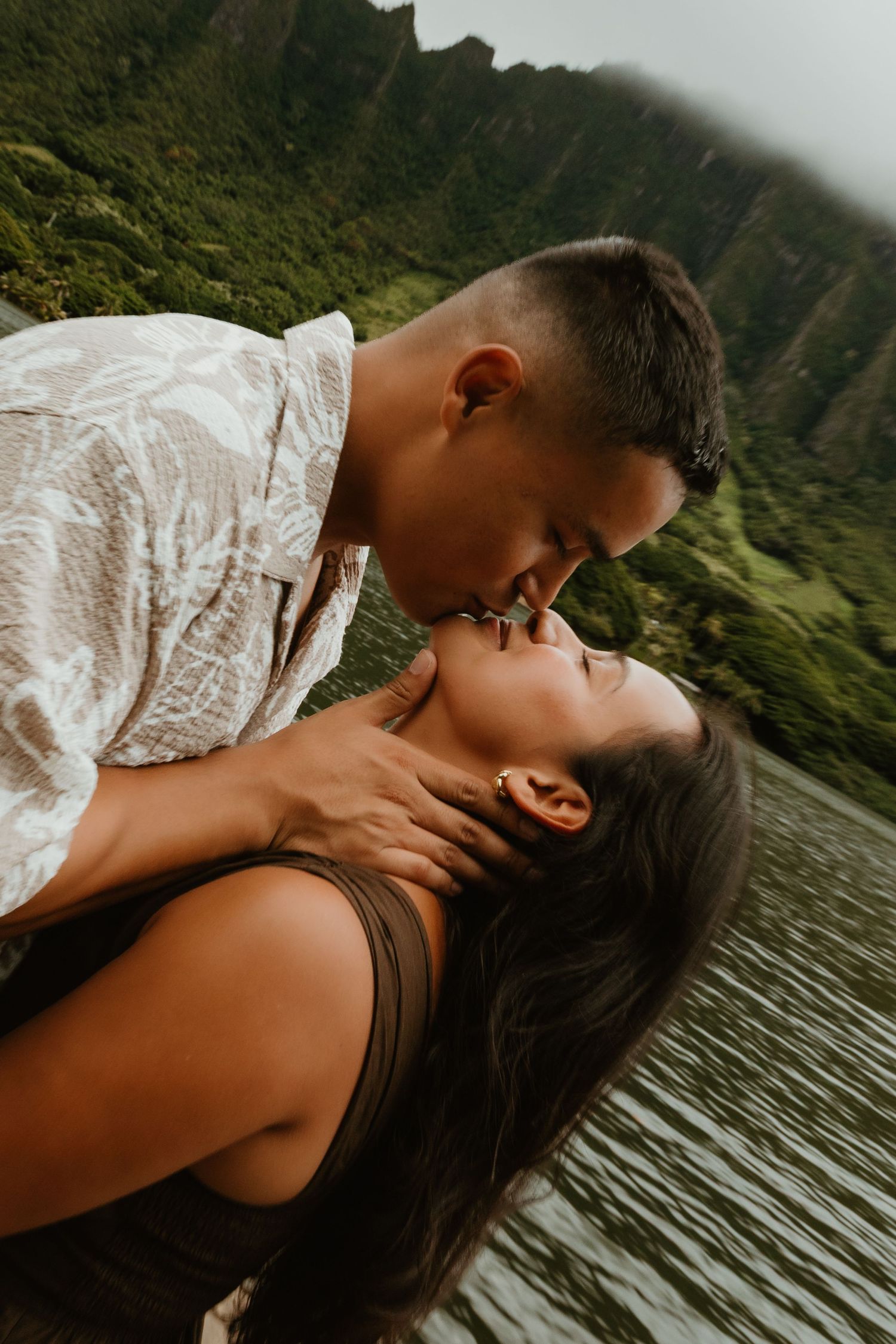 Couple sharing a romantic kiss by a serene lake with lush green mountains in Hawaii.