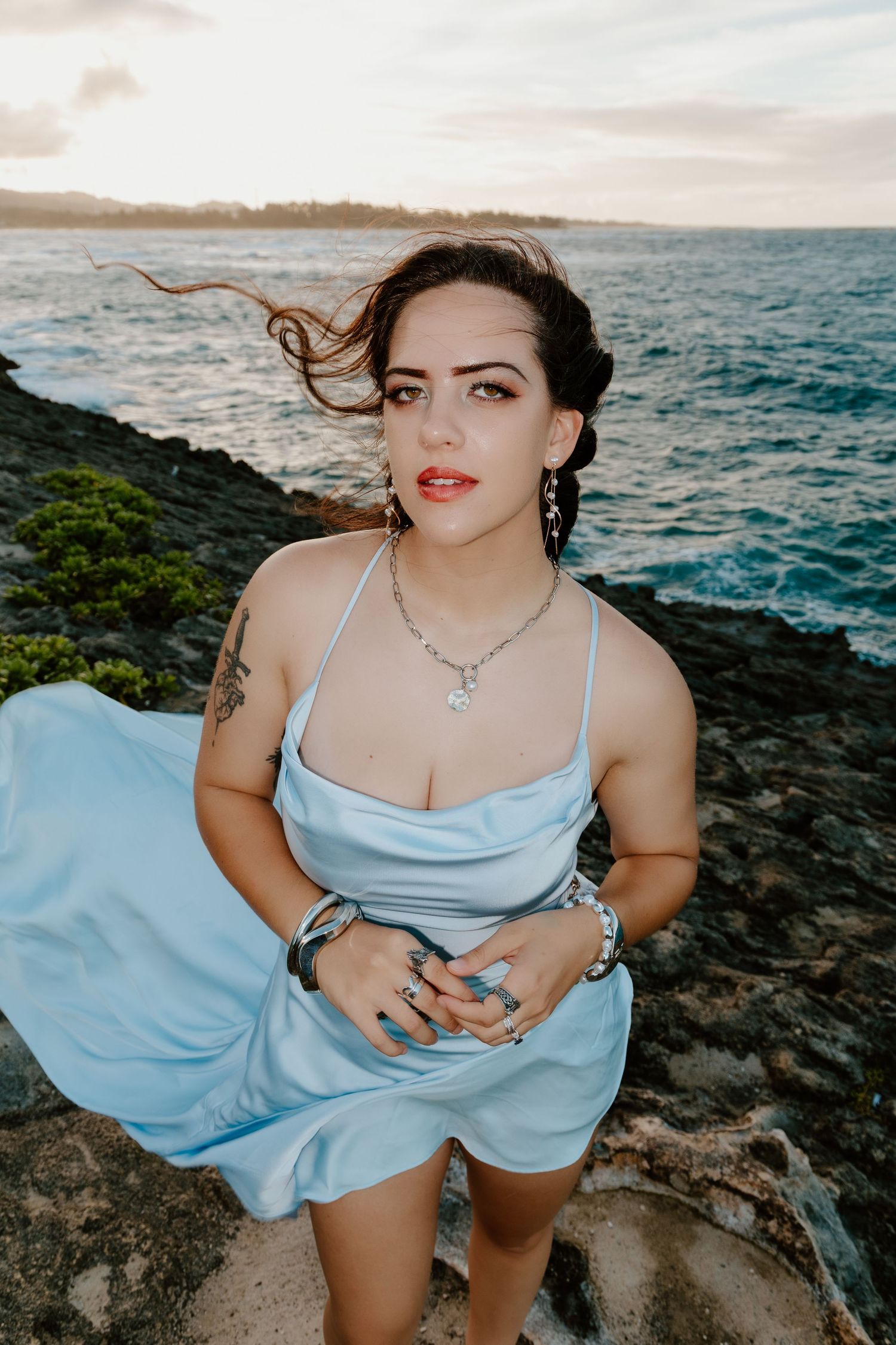 Woman in light blue satin dress poses on rocky shoreline at sunset, hair blowing in the wind.