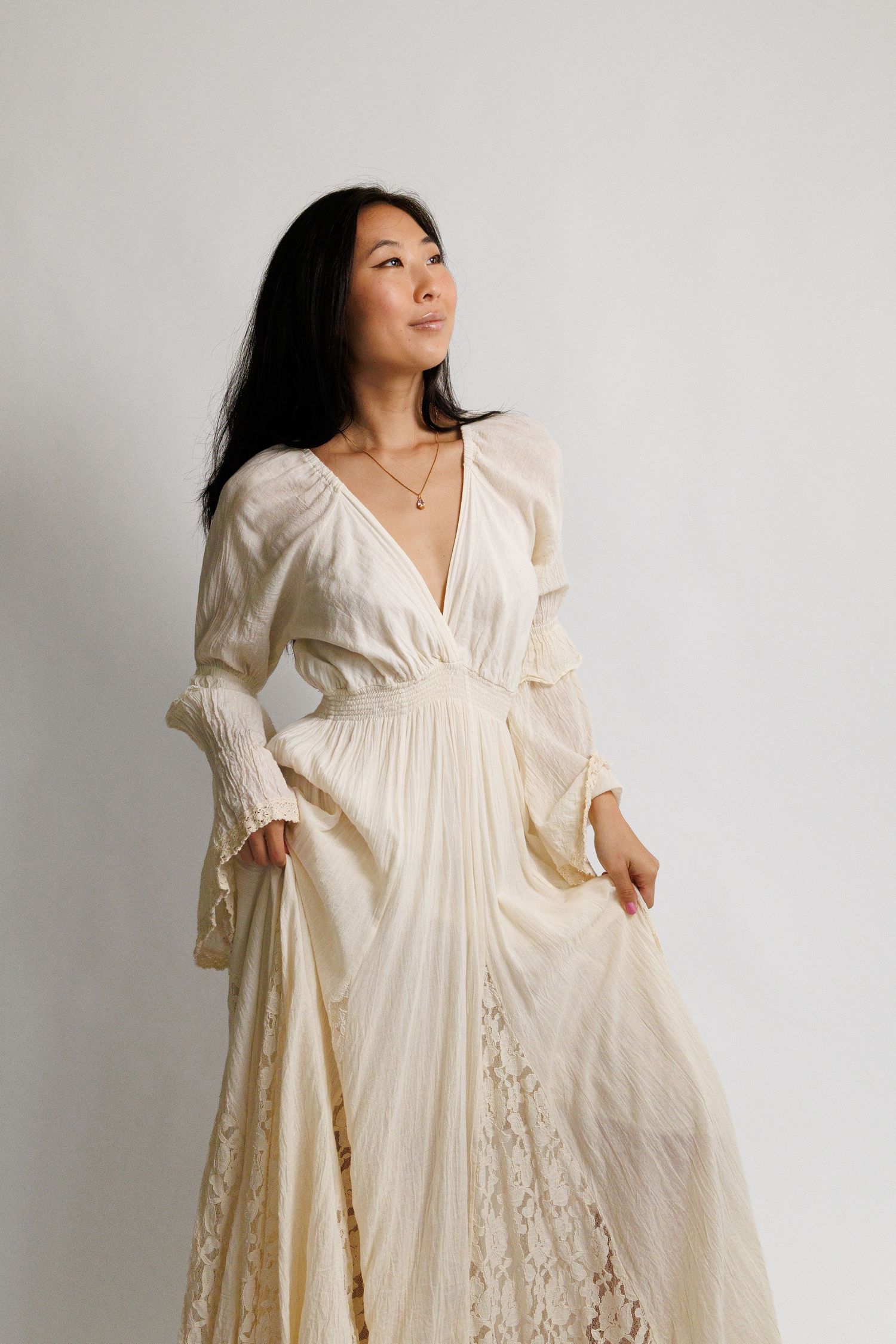 Woman in elegant cream lace long-sleeve maxi dress posing gracefully against white background.