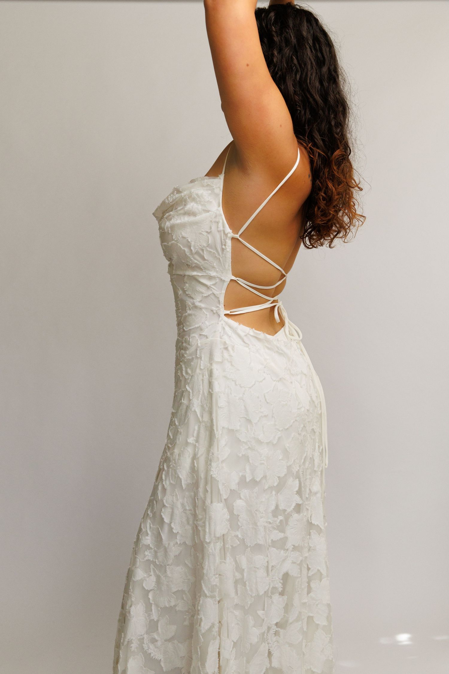 Woman in white lace backless dress with criss-cross straps, posing with arm raised, showcasing elegant open back design.