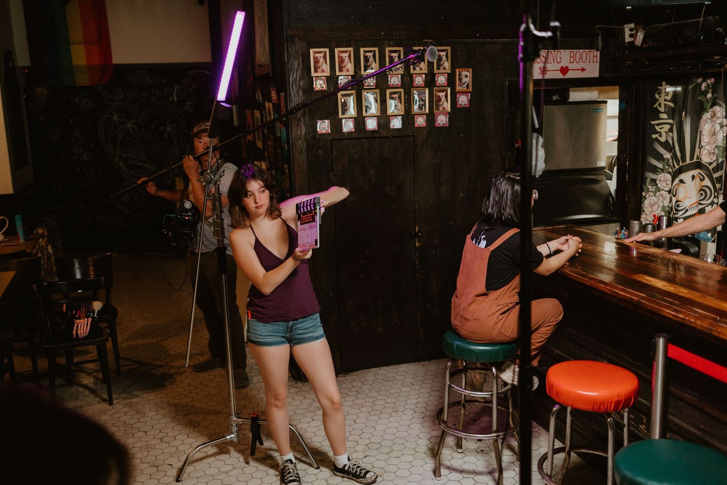Woman poses with pink toy gun under neon light in a decorated bar with colorful stools and photography equipment.