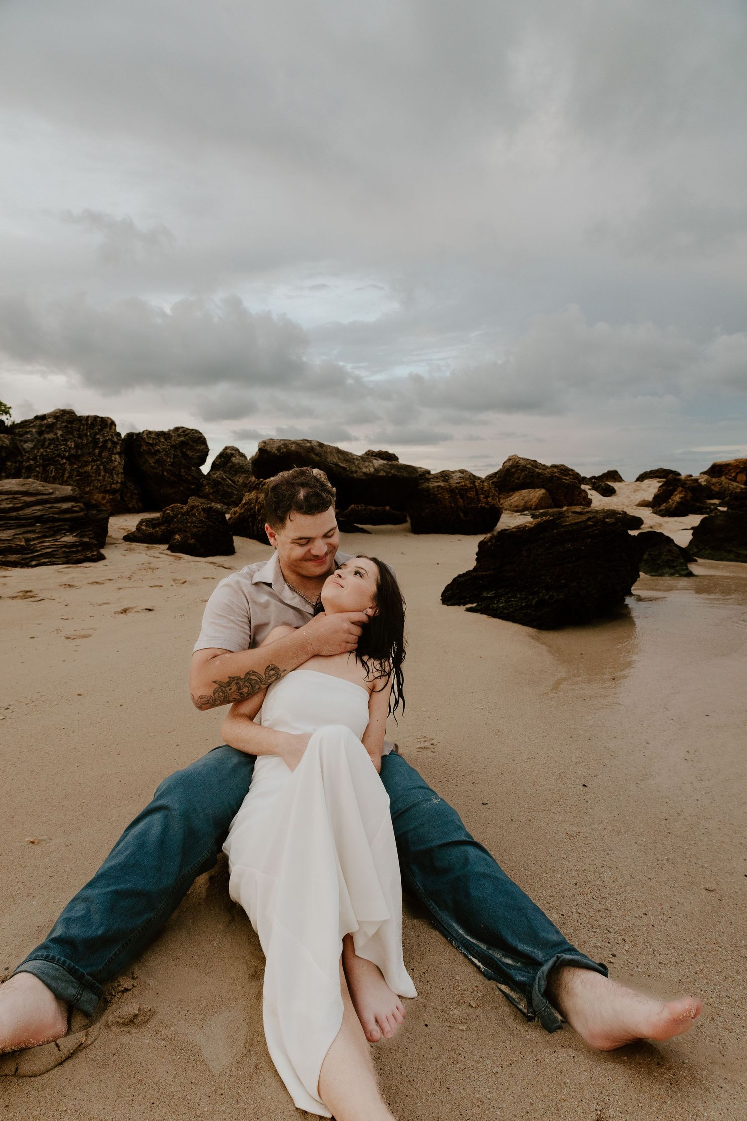 Couple sitting together on sandy beach near rocks, woman in white dress leaning back into mans arms laughing.