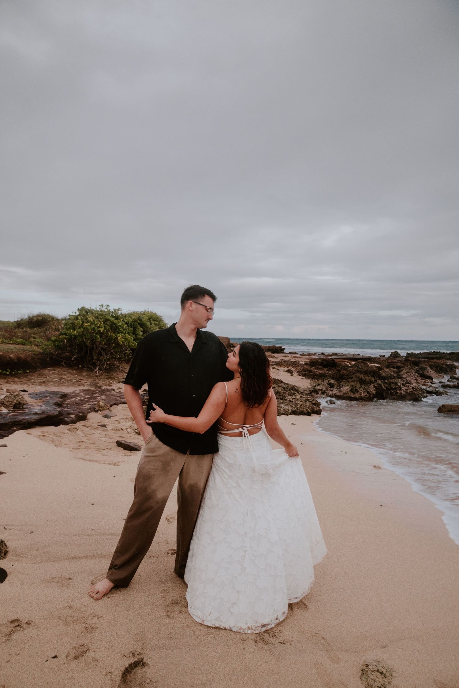 A wedding couple stands together on a sandy beach gazing at each other with rocky coastline behind them.