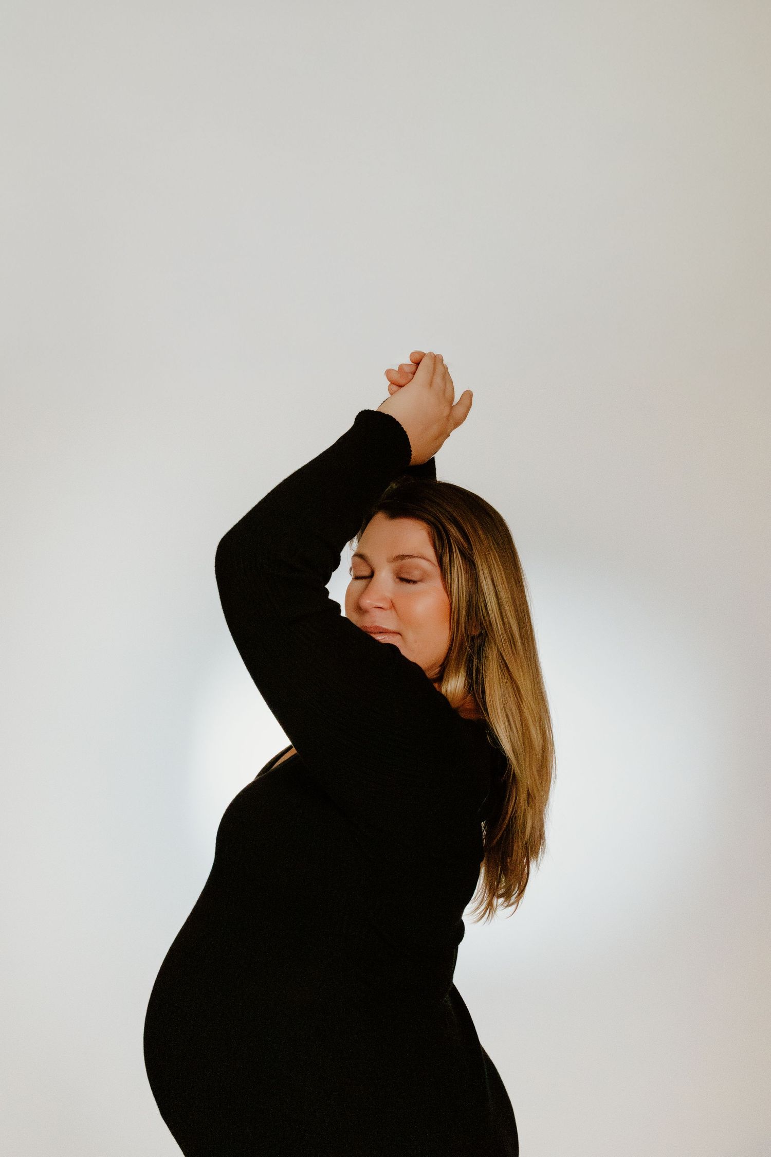 Pregnant woman in black dress posing with arms raised overhead, eyes closed, against white background.